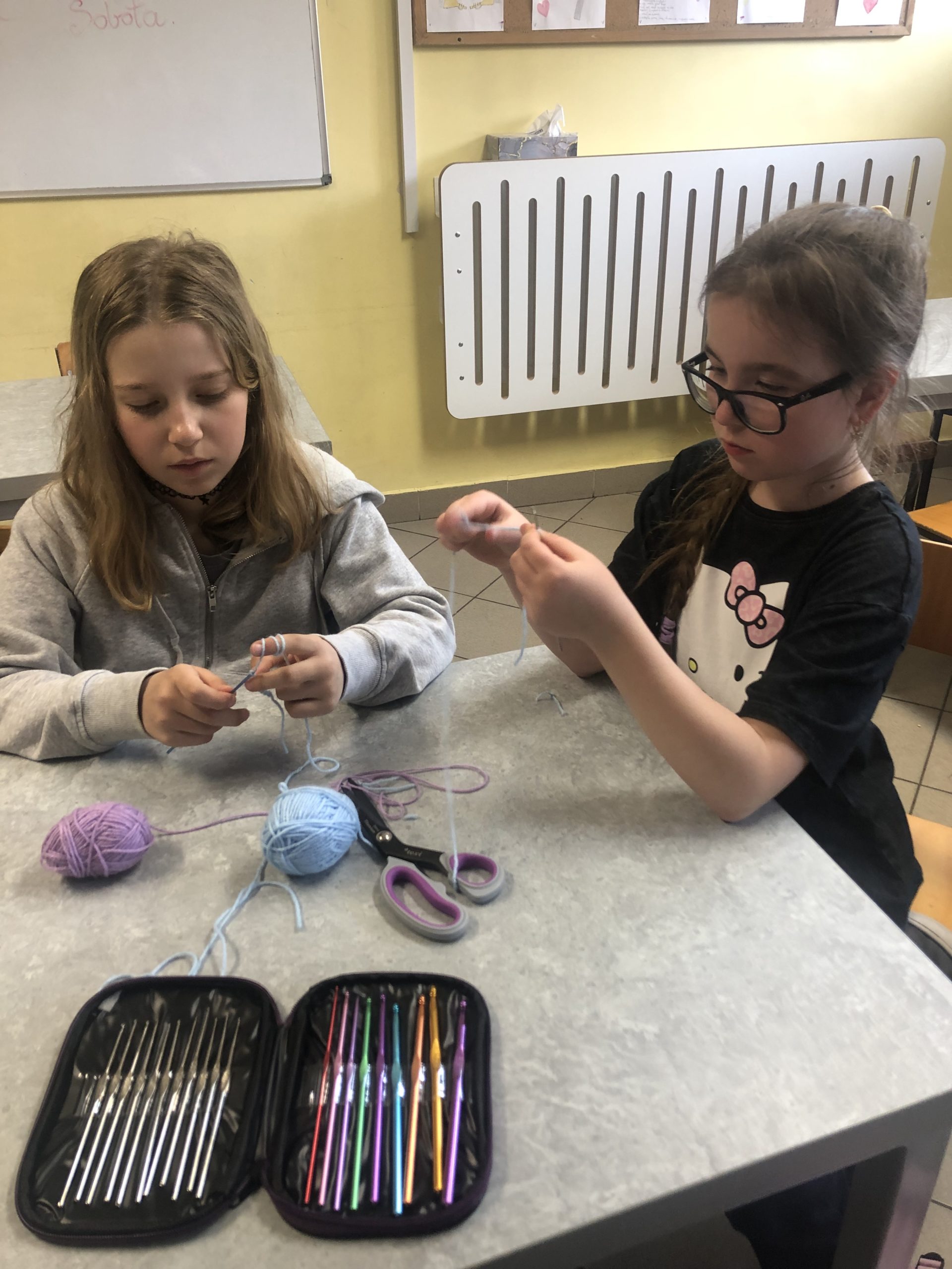 Dwie dziewczynki siedzą przy stoliku na którym leży komplet szydełek i włóczka.