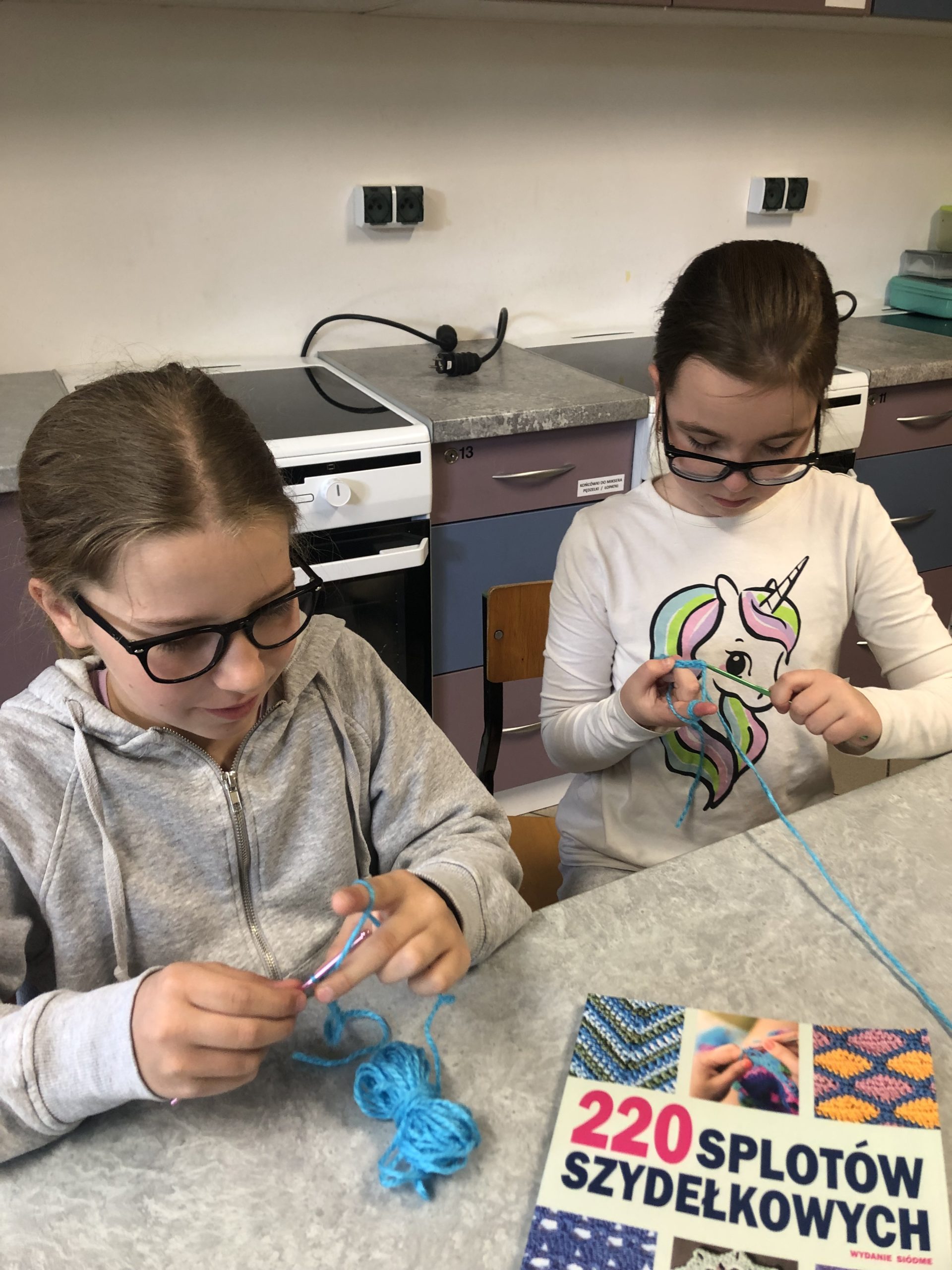 Dwie dziewczynki siedzą przy stole w rękach trzymają szydełko i włóczkę.