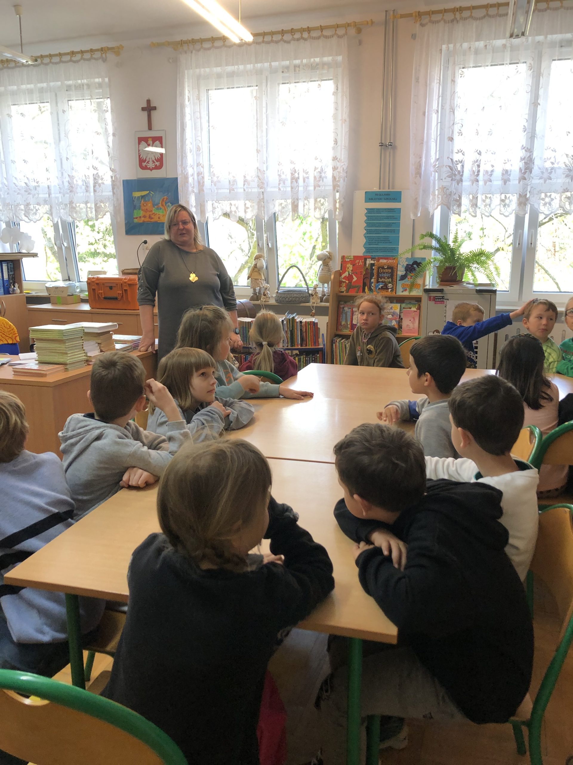 Grupa dzieci siedzi przy stolikach, z lewej strony stoi pani bibliotekarka.