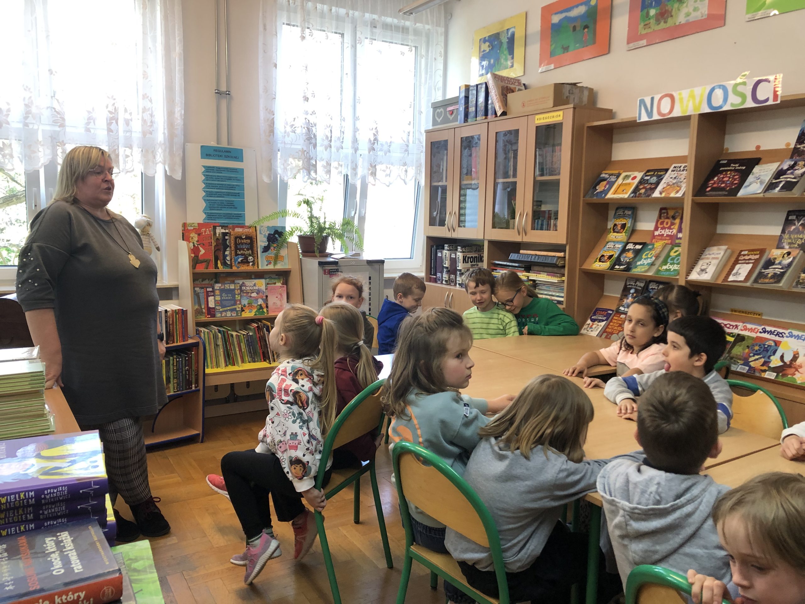 Grupa uczniów siedzi przy stolikach , za nimi przy oknie stoi pani bibliotekarka.