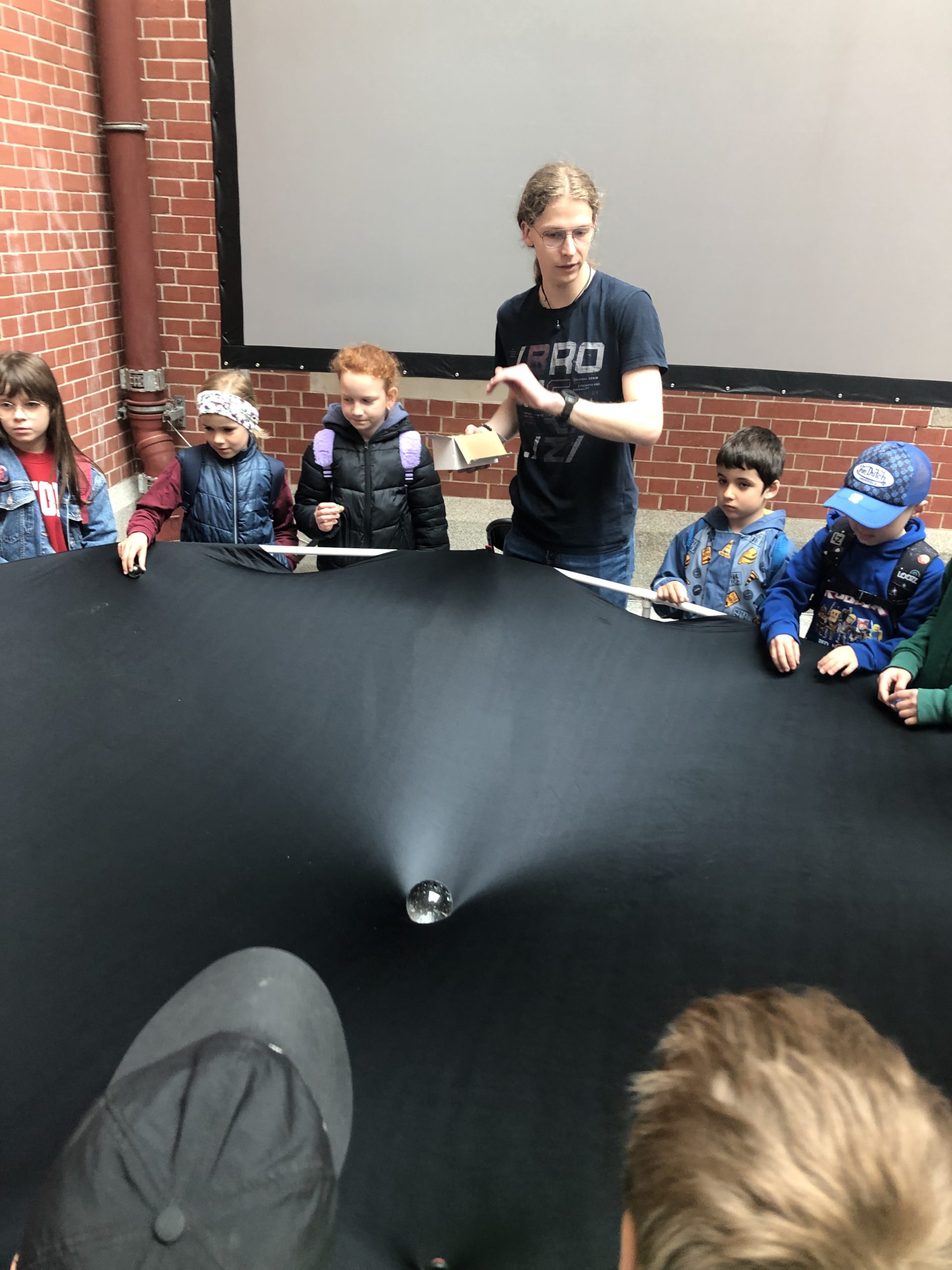 Grupa uczniów i jeden student stoi wokół rozpiętej czarnej płachty zakrzywionej przez kuleę umieszczoną na środku.