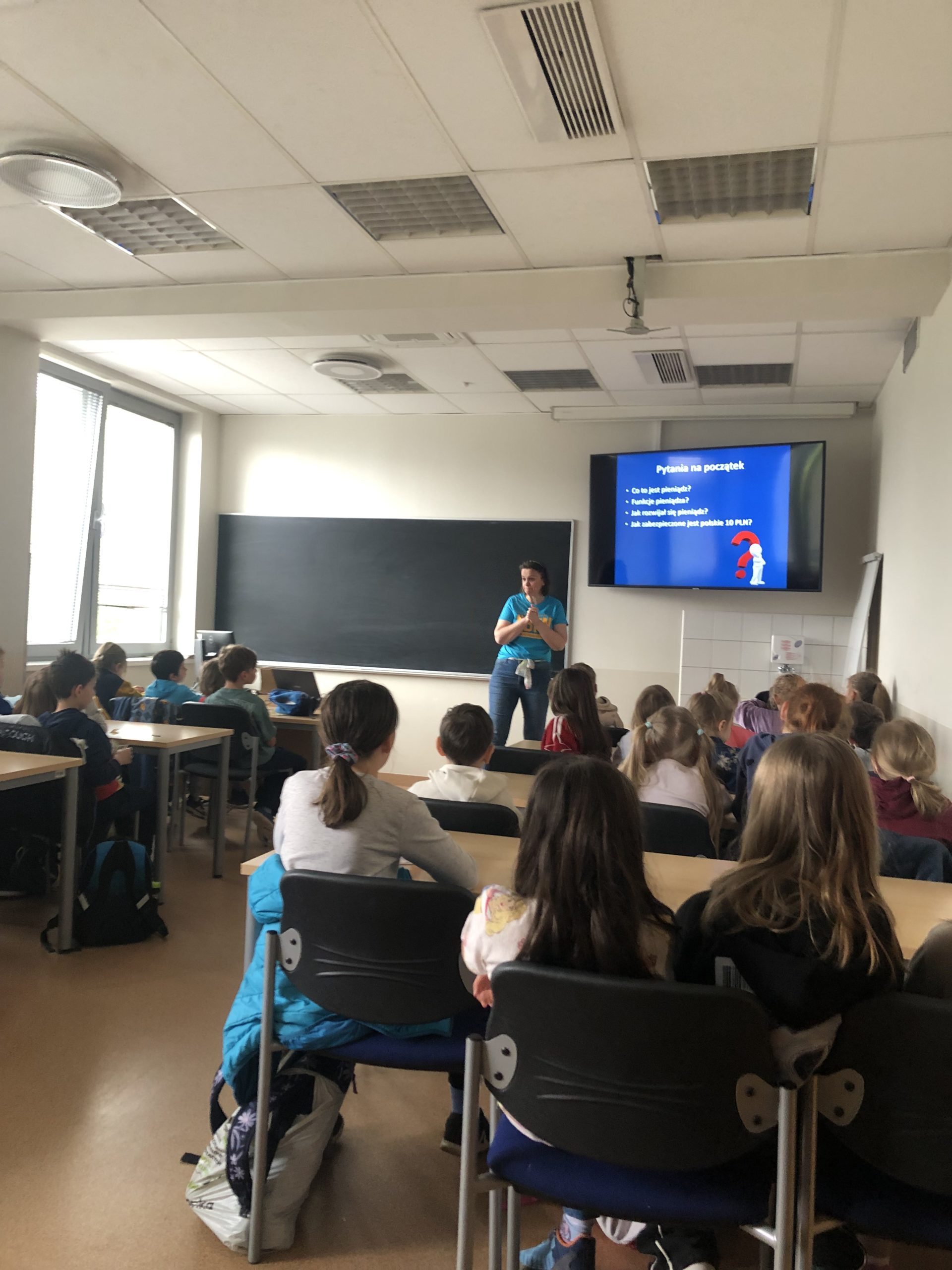 Grupa dzieci siedzi w ławkach w sali, na środku stoi pani, z prawej strony jest ogromny monitor.