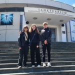 Trójka młodych osób - 2 dziewczyny i chłopak, stoi na schodach przed budynkiem Sanktuarium Najświętszej Maryi Panny Wspomożenia Wiernych. Pogoda jest słoneczna. Dwie dziewczyny chwytają się za ramiona.