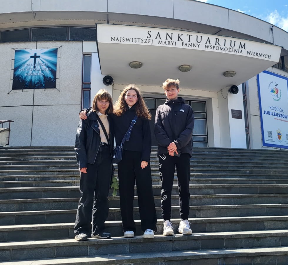 Trójka młodych osób - 2 dziewczyny i chłopak, stoi na schodach przed budynkiem Sanktuarium Najświętszej Maryi Panny Wspomożenia Wiernych. Pogoda jest słoneczna. Dwie dziewczyny chwytają się za ramiona.