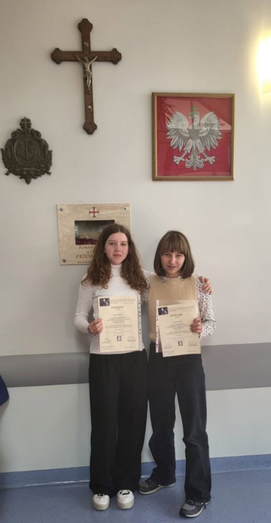 Dwie dziewczyny stoją w budynku pod ścianą, trzymając w rękach dyplomy. Za nimi widać krzyż i godło Polski oraz. Otoczenie sugeruje, że zdjęcie zostało zrobione w szkole.