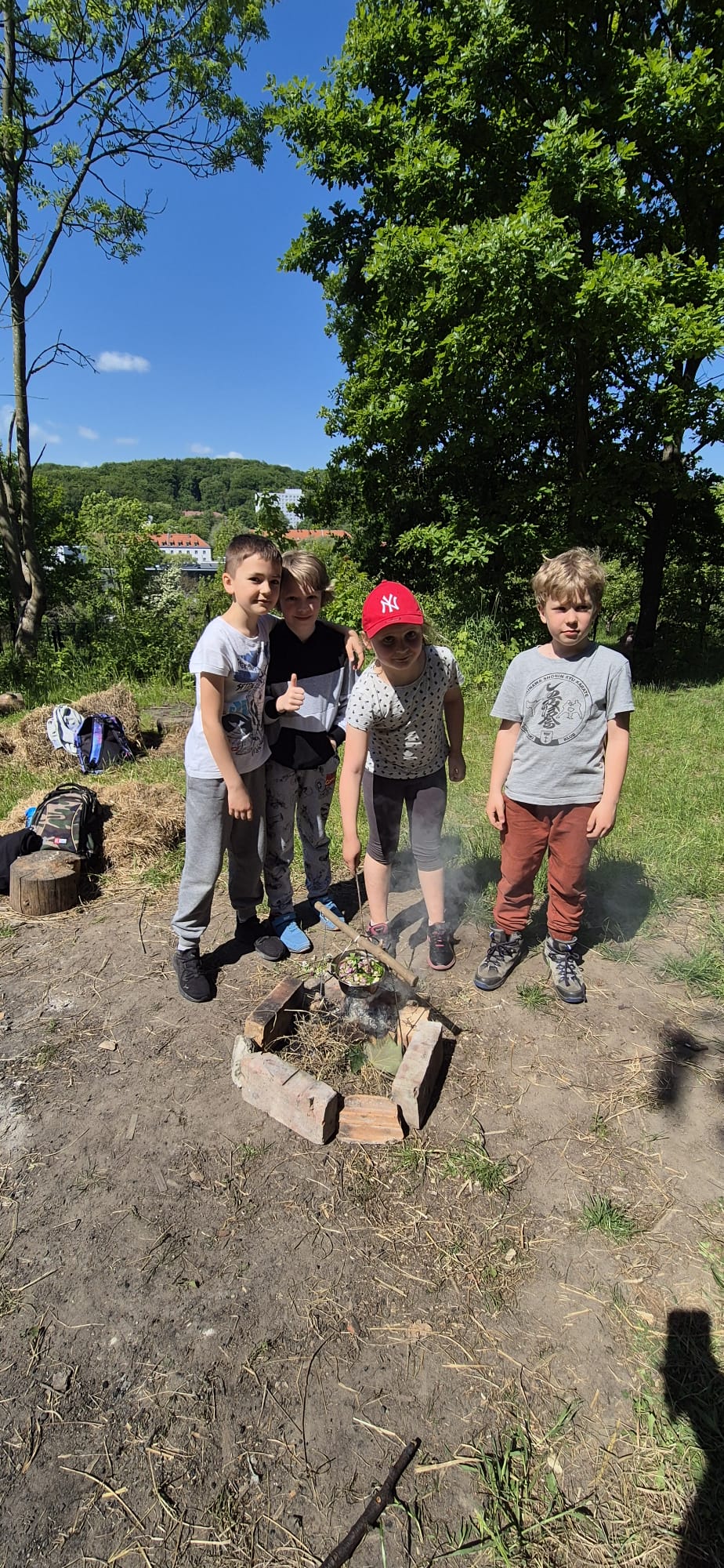 Dzieci stoją i obok siebie przy ognisku, gotują herbatkę kwiatową.