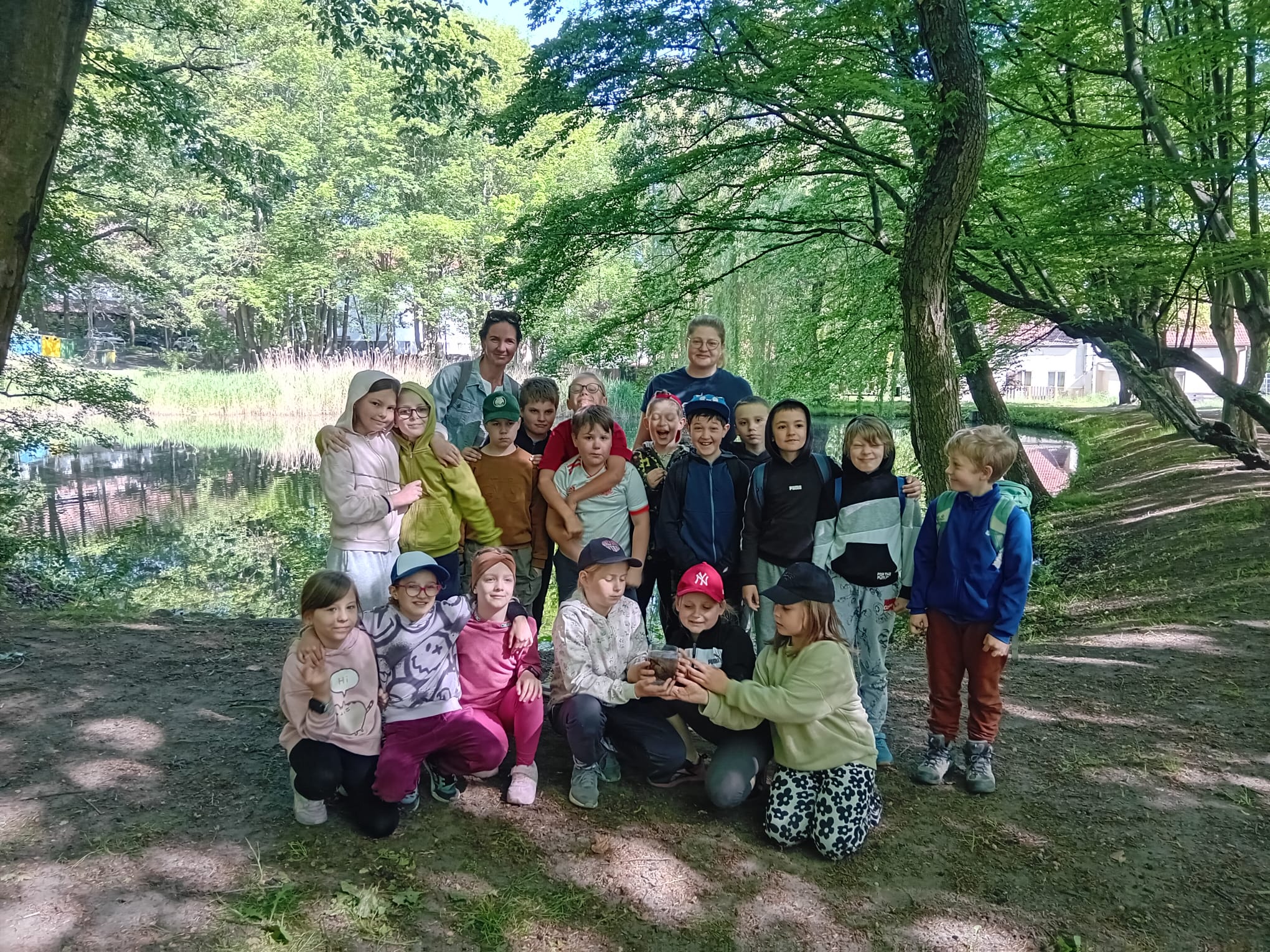 Grupa 18 dzieci stoi z dwoma Panią w lesie. Dzieci stoją obok siebie. Za dziećmi są drzewa.