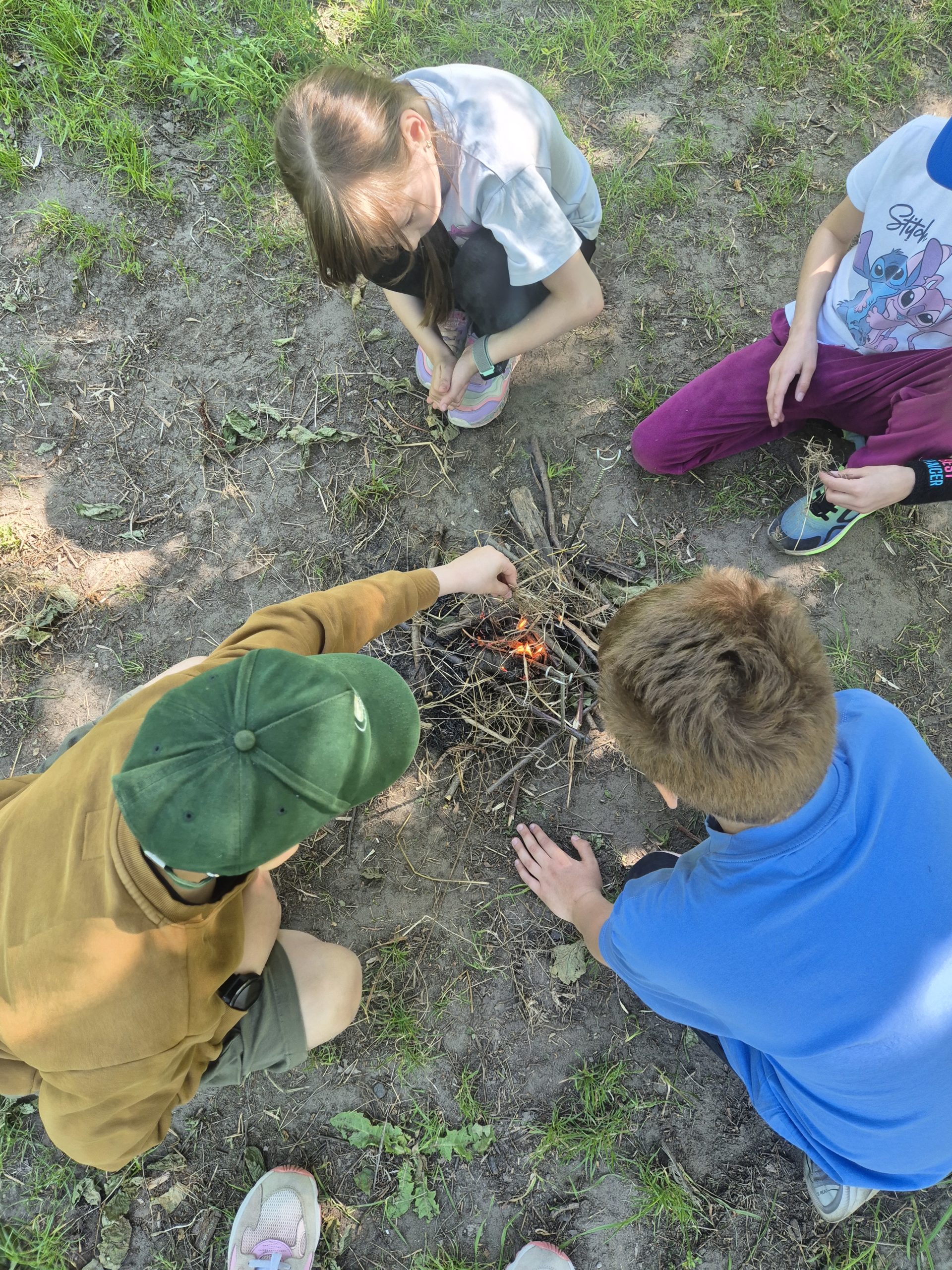 Rozpalone ognisko, które dosyć słabo widać. Są widoczne głowy dwójki dzieci. Dzieci to ognisko rozpalały samodzielnie.