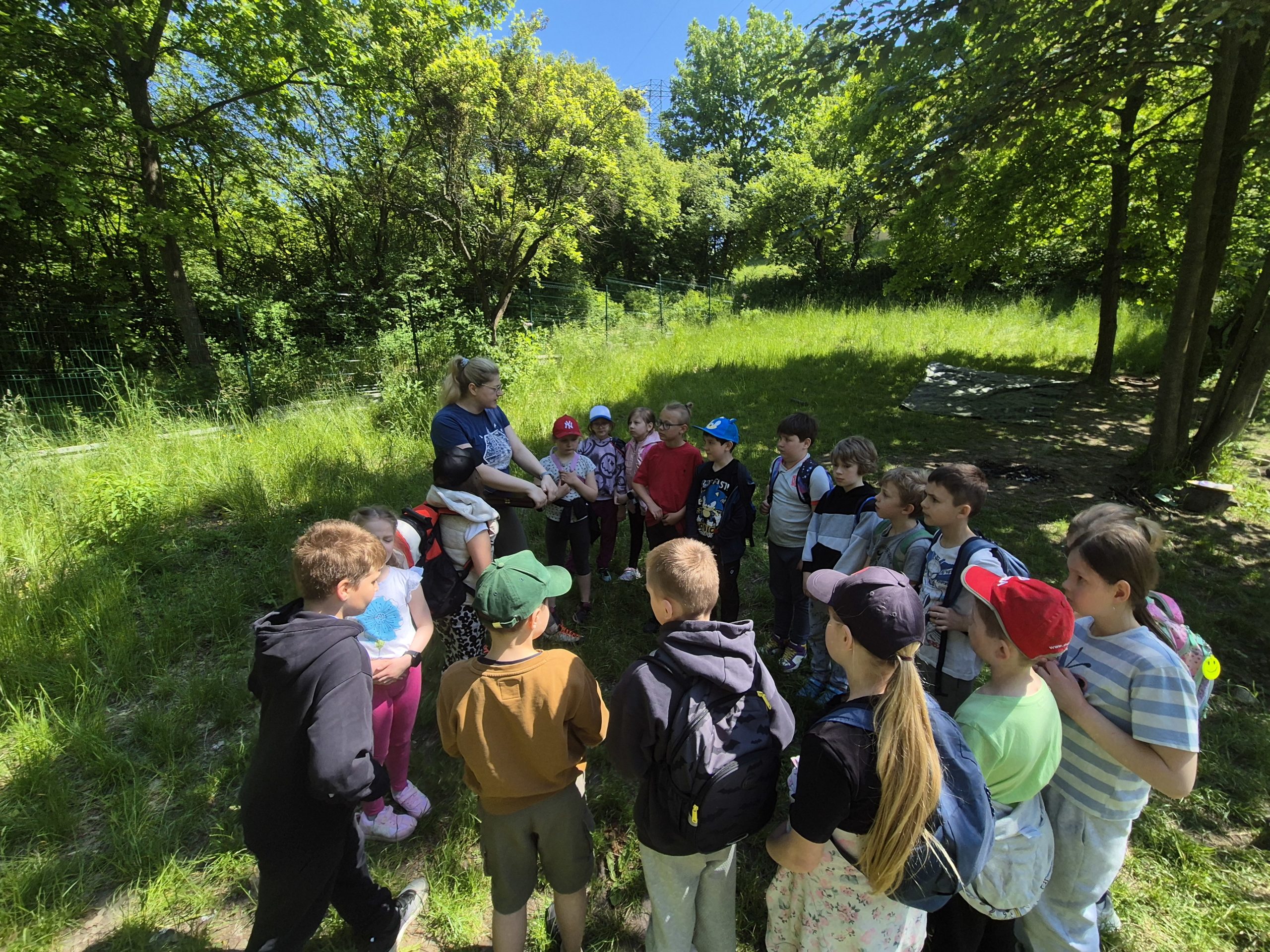  kole stoją dzieci. Jest ich osiemnaście. Jest z nimi Pani która trzyma w ręku pudełko a w nim odznaki dla dzieci.