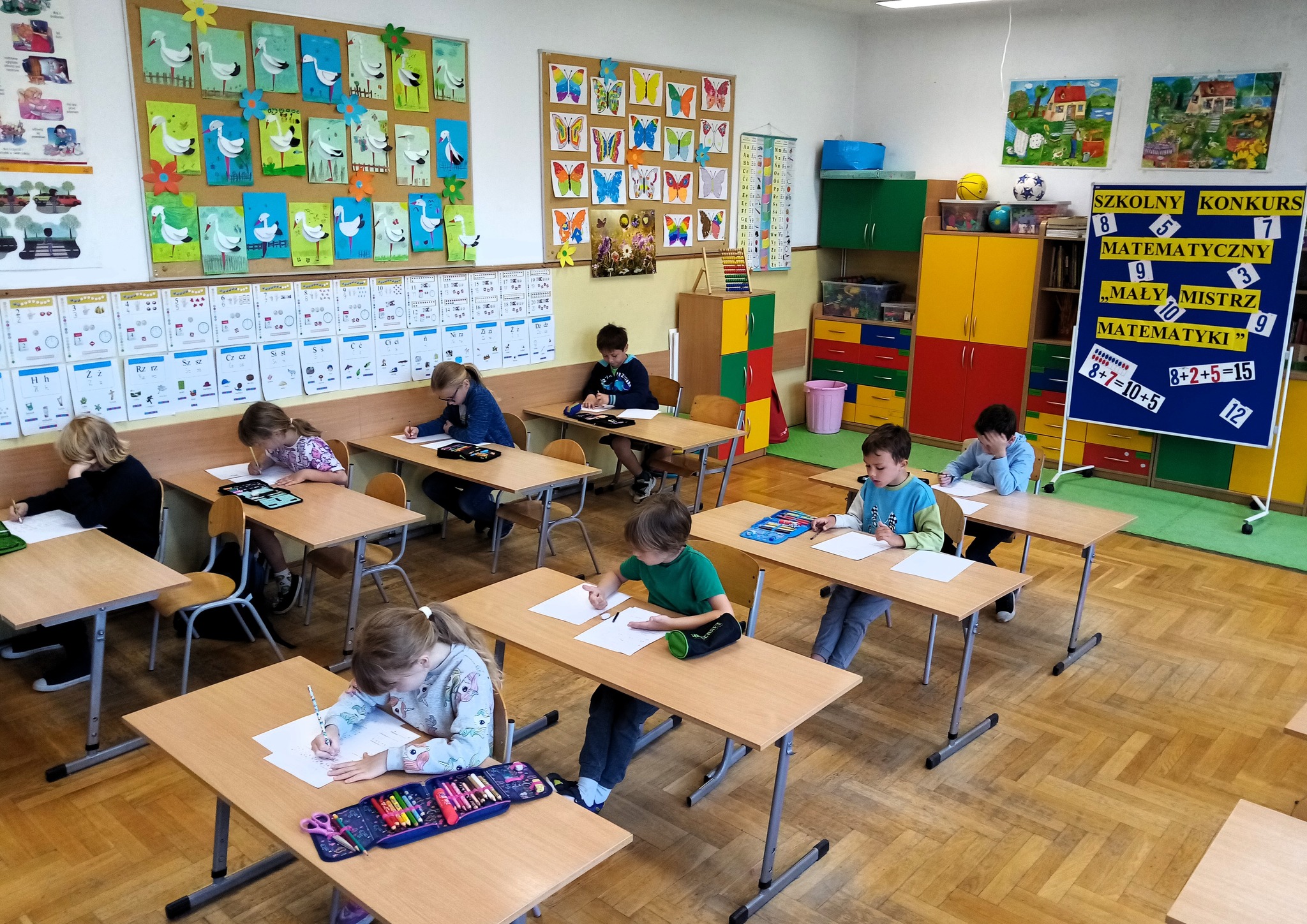8 dzieci siedzące przy oddzielnych ławkach, rozwiązujące zadania matematyczne. W tle kolorowe dekoracje z motylami, ptakami oraz tablica informująca o konkursie matematycznym z napisem: 'Szkolny Konkurs Matematyczny - Mały Mistrz Matematyki'. W tle prace plastyczne, zabawki, szafki.