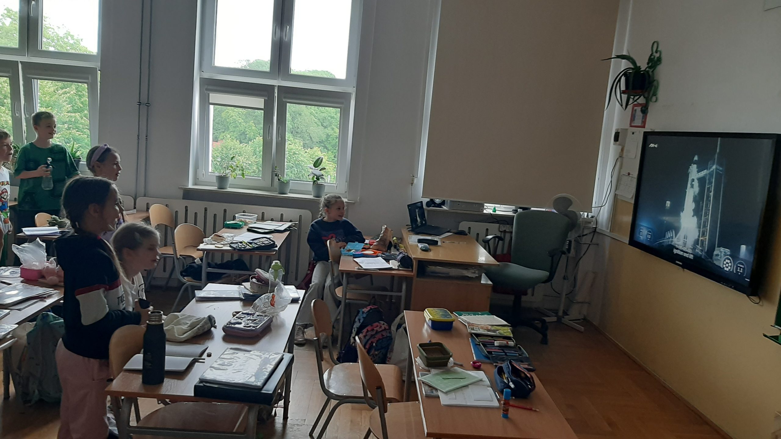 Grupa dzieci w sali lekcyjnej. Po prawej ekran, na którym widać rakietę przygotowującą się do startu. Jest odpalana. W sali są dwa rzędy ławek ustawione frontem do ekranu. Na ławkach leżą przybory szkolne.W ławkach siedzą dwie dziewczynki. Pozostałe cztery osoby stoją. Wszyscy patrzą na ekran.