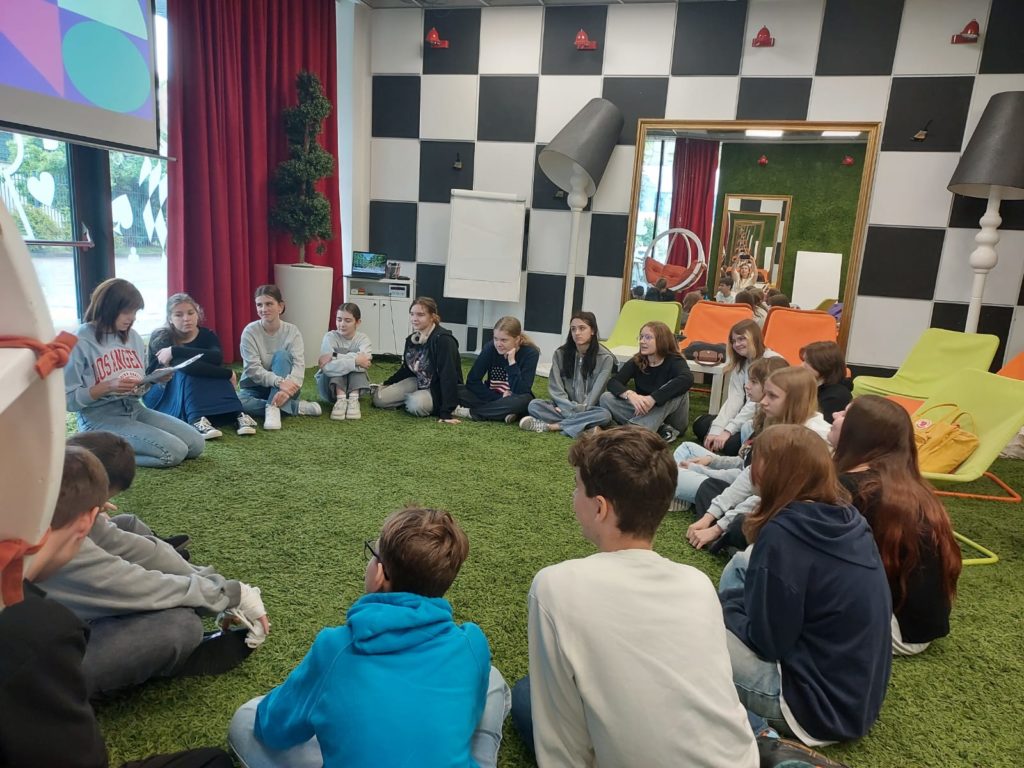 Uczniowie klasy 7b siedzą w kręgu na zielonym dywanie, który przypomina trawę. Znajdują się w kolorowym i nowocześnie urządzonym pomieszczeniu z czarno-białą szachownicą na ścianie, dużym lustrem i nietypowymi, dużymi lampami stojącymi pod kątem. Po lewej stro-nie widoczne jest okno z czerwonymi zasłonami, przez które widać serduszka przyklejone na szybie. Prowadząca zajęcia pani w bluzie z napisem „Los Angeles” trzyma zeszyt i mówi do reszty grupy.