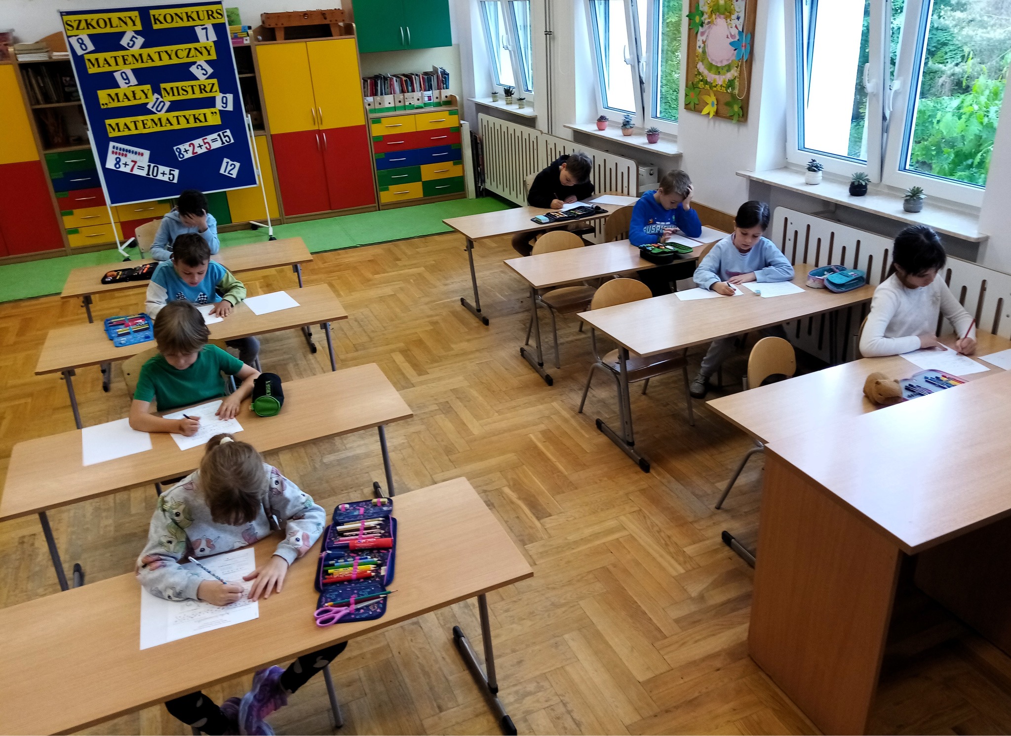8 dzieci siedzące przy oddzielnych ławkach, rozwiązujące zadania matematyczne. W tle tablica informująca o konkursie matematycznym z napisem: 'Szkolny Konkurs Matematyczny - Mały Mistrz Matematyki'. 