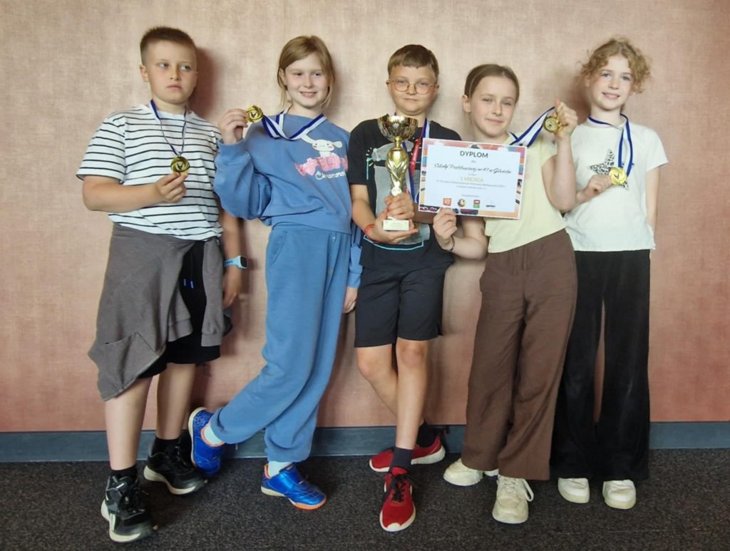 Pięcioro uczniów z klasy 3a stoi na tle różowej ściany, uśmiechniętych i dumnie prezentujących swoje osiągnięcie. Każde z nich ma na szyi złoty medal, a dziecko w środku trzyma puchar. Dziecko obok niego trzyma dyplom. Wszyscy są ubrani na sportowo.