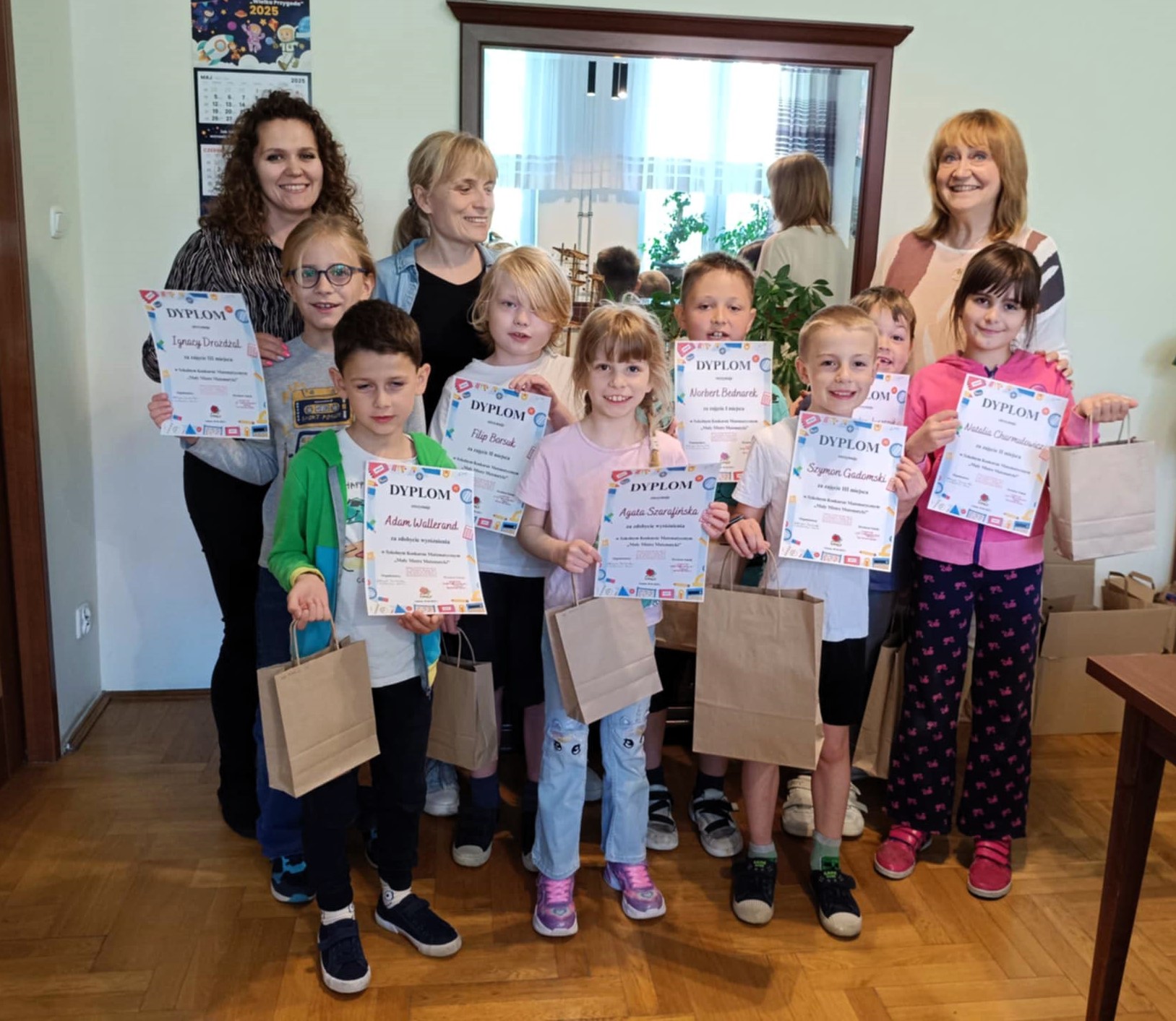 3 panie oraz grupa uśmiechniętych dzieci stoi w rzędzie razem z trzema nauczycielkami, trzymając dyplomy i papierowe torby z nagrodami. 