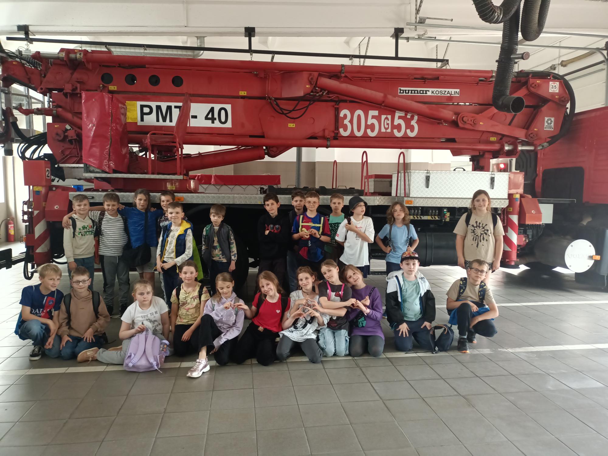 Dwudziestu pięciu uczniów pozuje w hali garażowej na tle dużego wozu strażackiego z podnośnikiem.  Dzieci ustawione są w dwóch rzędach. Pierwszy siedzi na podłodze, a drugi rząd stoi.