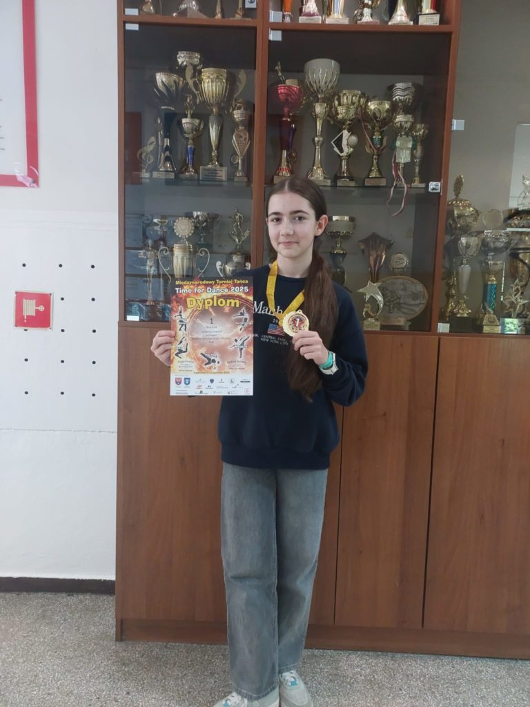 Uczennica klasy 7b Julianna Osasiuk stoi przed przeszkloną gablotą pełną pucharów. Dziew-czyna trzyma w jednej ręce dyplom, a w drugiej złoty medal zawieszony na żółtej wstążce. Na dy-plomie widnieje napis „Międzynarodowy Turniej Tańca – Time for Dance 2025” oraz informacja o zdobyciu 1. miejsca. Ubrana jest w ciemną bluzę i luźne, szare dżinsy, a na nogach ma sportowe buty.