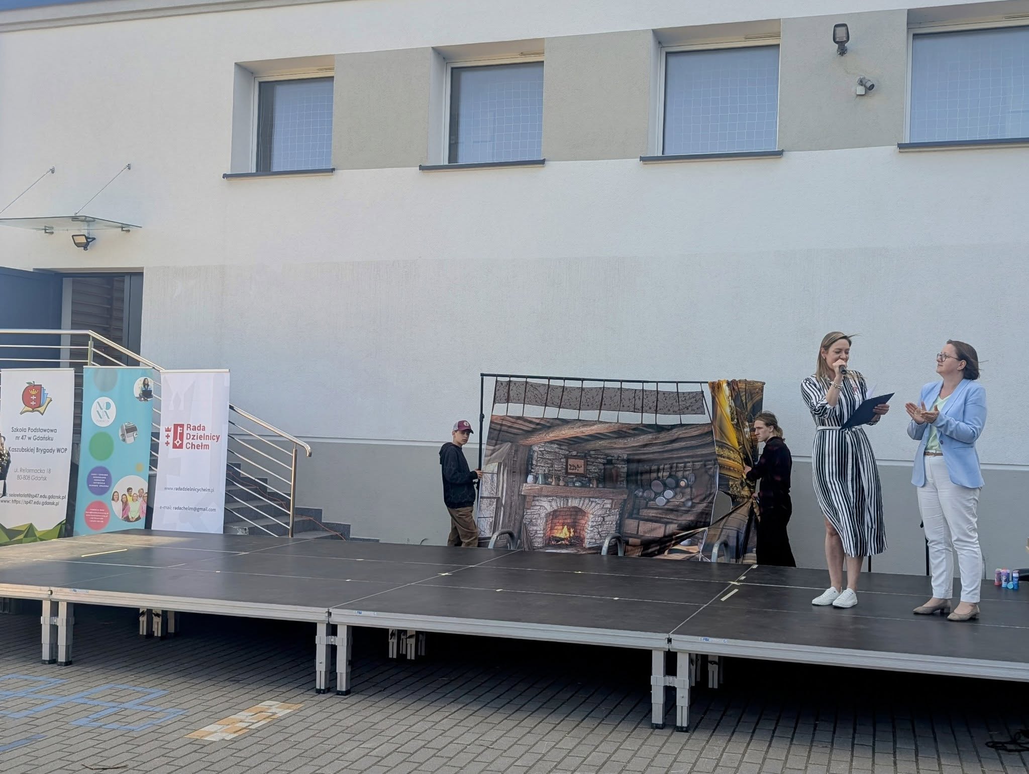 Na scenie stoją 2 dyrektorki szkoły, jedna z nich trzyma mikrofon. W tle budynek szkoły, po lewej stronie 3 banery sponsorów festynu