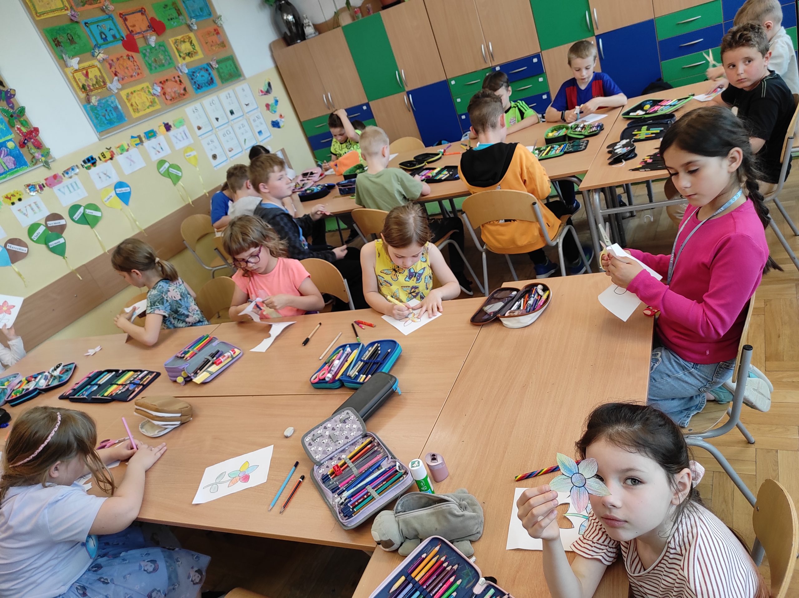 U. siedzą przy stołach, podzieleni na dwie grupy. Na stołach leżą piórniki. Dzieci trzymają w dłoniach kartki i nożyczki.