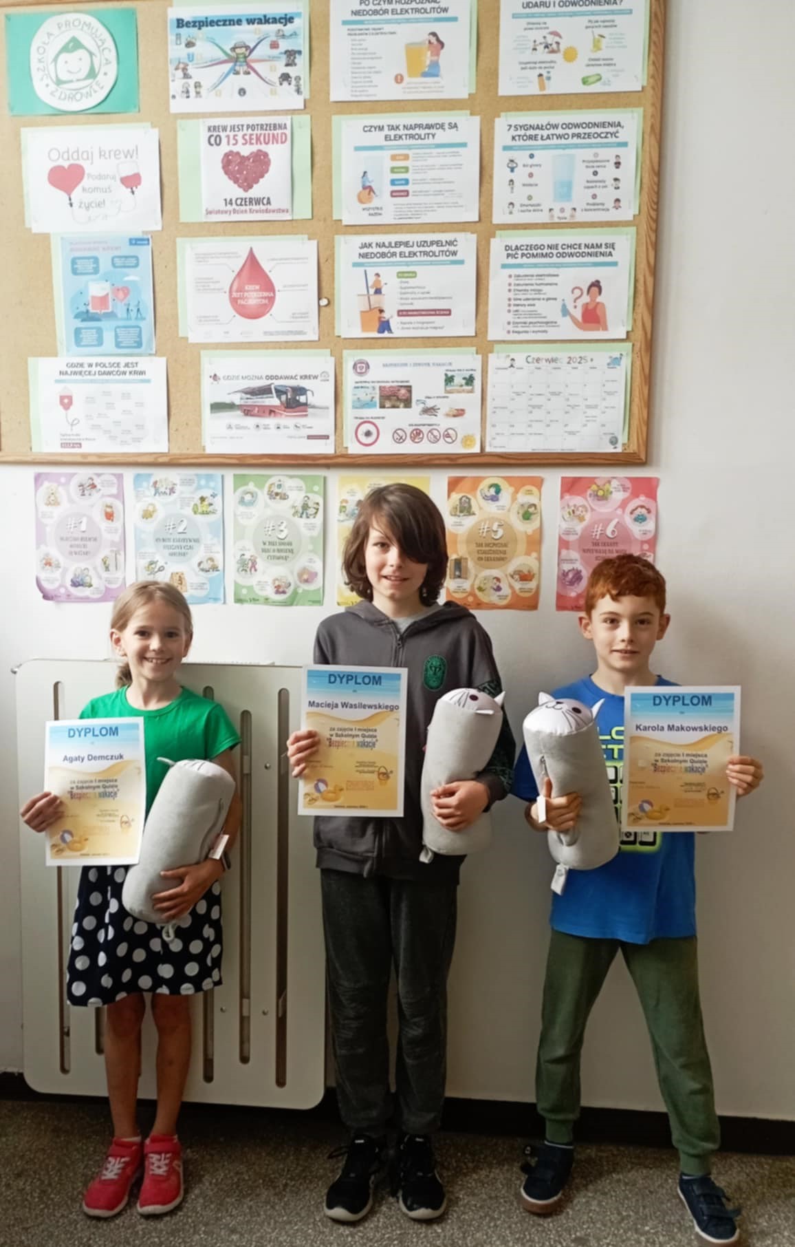 Na szkolnym korytarzu na tle tablicy z ogłoszeniami na temat zdrowia stoi dwóch uczniów i jedna uczennica klas młodszych. W rękach trzymają dyplomy i nagrody.