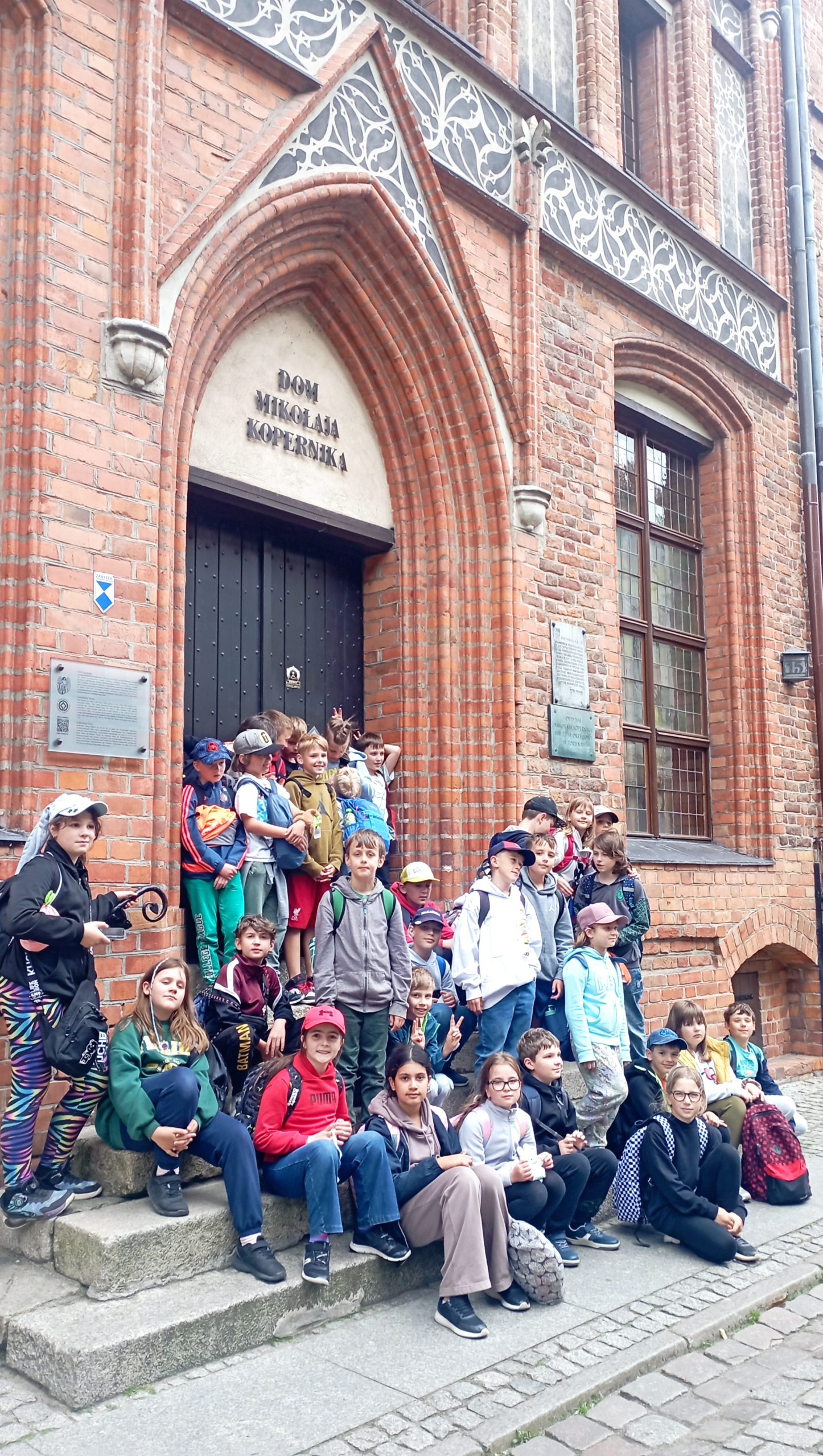 Grupa uśmiechniętych dzieci stoi przed fasadą zabytkowego domu z napisem DOM MIKOŁAJA  KOPERNIKA