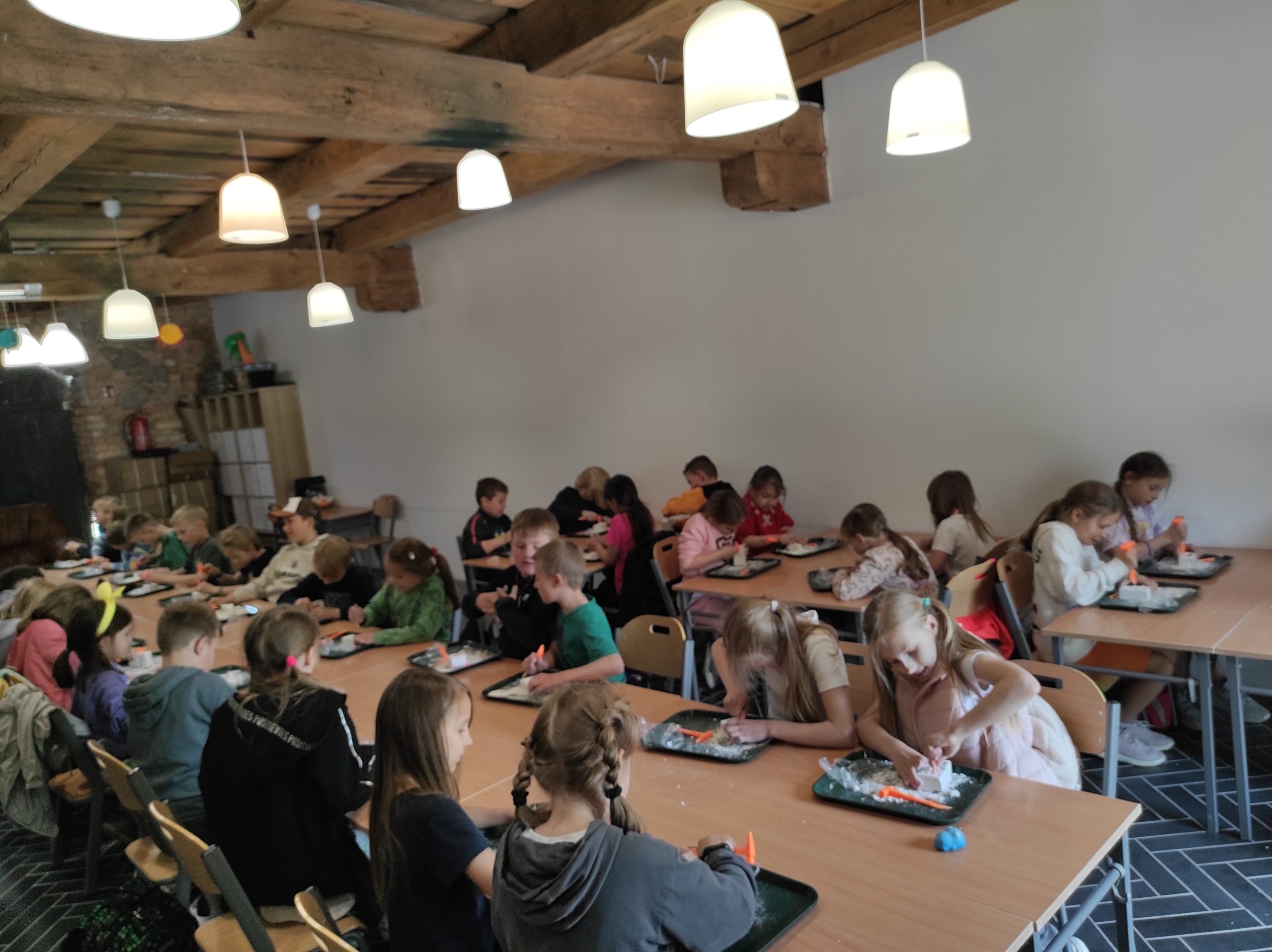 Dzieci siedzą przy zsuniętych ze sobą stolikach, twarzami do siebie. Przed każdym z uczniów leży taca, młoteczek zabawkowy. Dzieci są skupione na pracy.