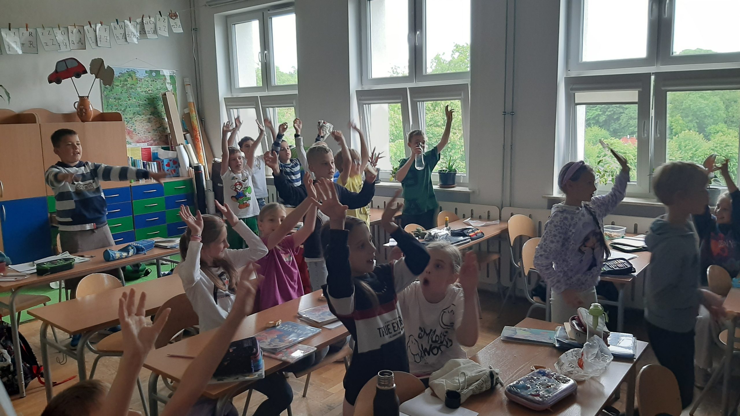 Grupa dzieci w klasie. Ławki ustawione w dwóch rzędach. Po lewej fragment regału i mapa Polski. W tle okna. Większość dzieci stoi, kilkoro siedzi.Wszyscy w geście radości podnoszą ręce do góry.