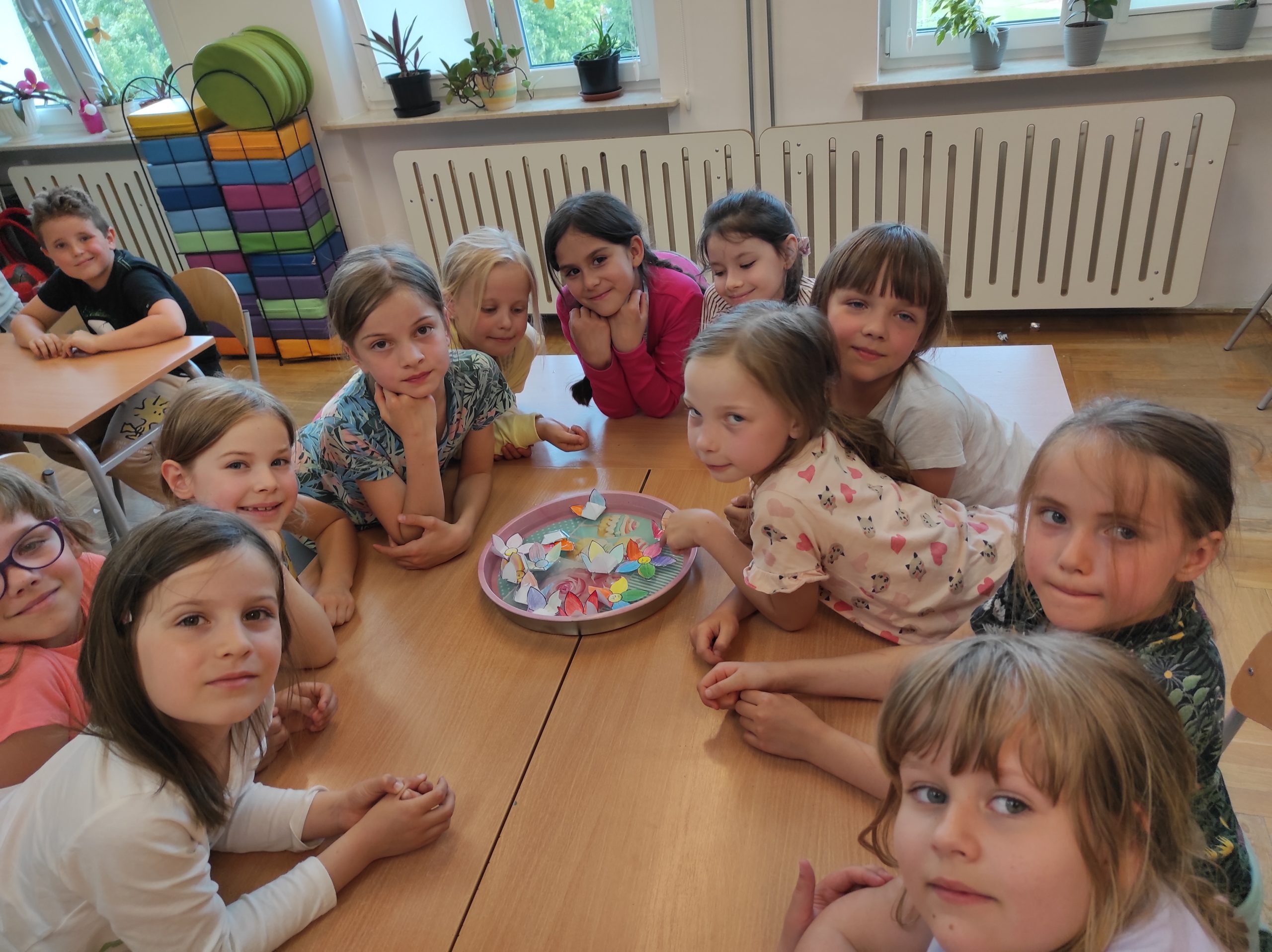 10 dziewczynek siedzi wokoło złączonych stołów. Na środku leży taca, a w niej widoczne są kwiaty z papieru. Kilka z nich jest częściowo zwinięta, reszta rozwinięta. W tle elementy sali.