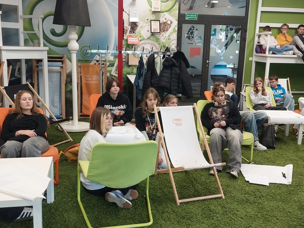 Uczniowie klasy 7b uczestniczą w zajęciach w nowocześnie urządzonym pomieszczeniu z zie-loną, sztuczną trawą na podłodze. Młodzież siedzi na kolorowych fotelach, leżakach i pufach, niektórzy na podłodze, pracując w mniejszych grupach. Przed nimi leżą duże arkusze papieru i przybory do pisania. Uczniowie pracują nad wspólnymi projektami i robią burzę mózgów. W tle widać szatnię z kurtkami, przeszklone drzwi oraz nowoczesną aranżację ścian z grafikami i inspirującymi hasłami.