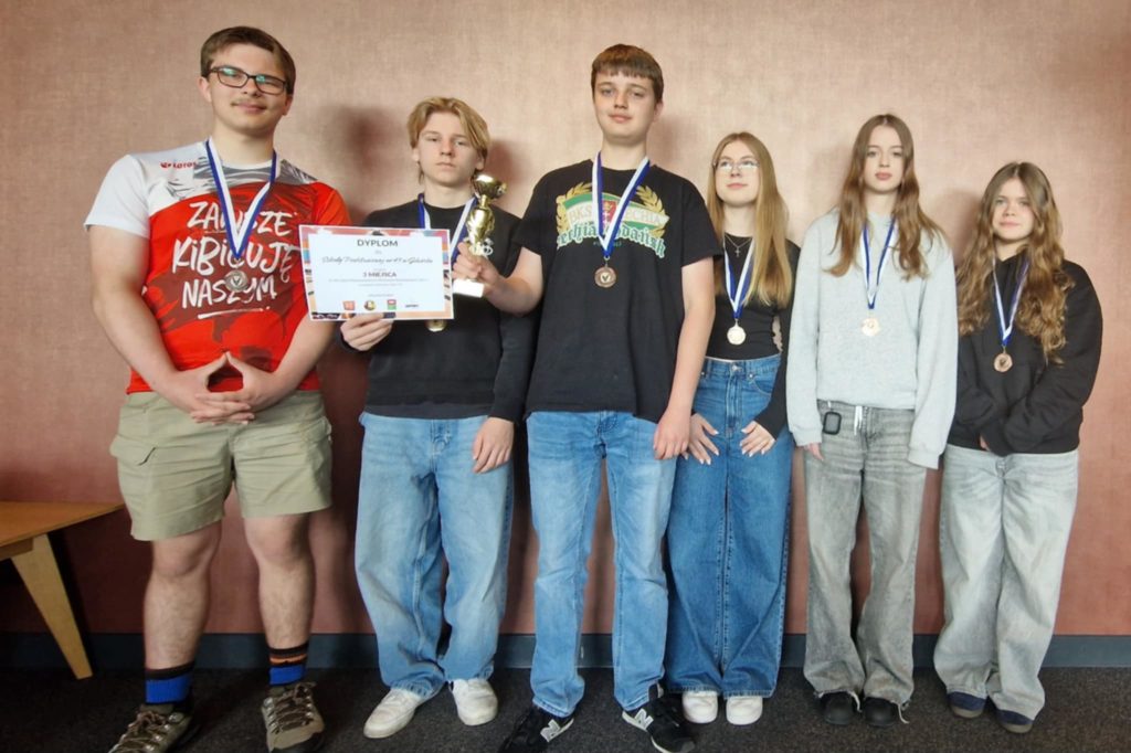 Sześcioro uczniów z klasy 8a stoi w szeregu na tle różowej ściany. Wszyscy mają na szyi brą-zowe medale i uśmiechają się . Chłopiec, który stoi w środku trzyma puchar, a obok niej druga osoba prezentuje dyplom. Uczniowie są ubrani w codzienne, wygodne ubrania.