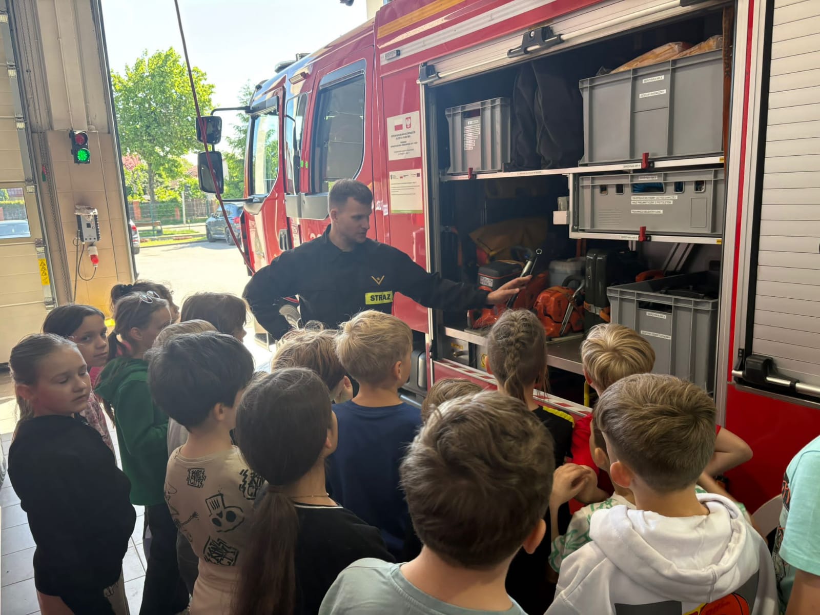 Grupa uczniów stoi przed otwartym wozem strażackim zaparkowanym w hali garażowej. Strażak pokazuje dzieciom wyposażenie wozu.