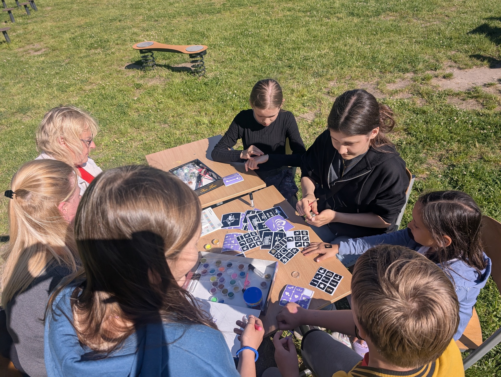na trawie przy stolikach siedzą 2 kobiety oraz 5 dzieci. Na stole rozłożone są tatuaże i kolorowe farbki.