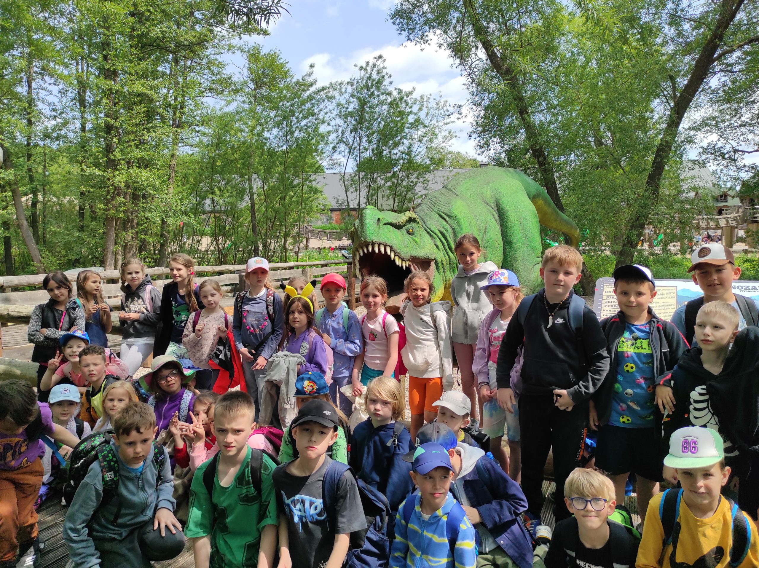  Zdjęcie w plenerze. 40 dzieci, cześć stoi, część siedzi. W tle duża makieta dinozaura i zieleń.