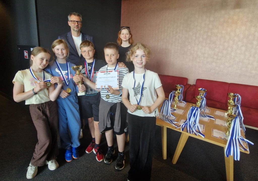 Pięcioro uczniów oraz dwie osoby dorosłe – mężczyzna i kobieta – stoją razem w grupie. Dzieci trzymają złote medale, puchar oraz dyplom z napisem I MIEJSCE. W tle, na stole po prawej stro-nie, widoczne są kolejne puchary, medale i dyplomy.