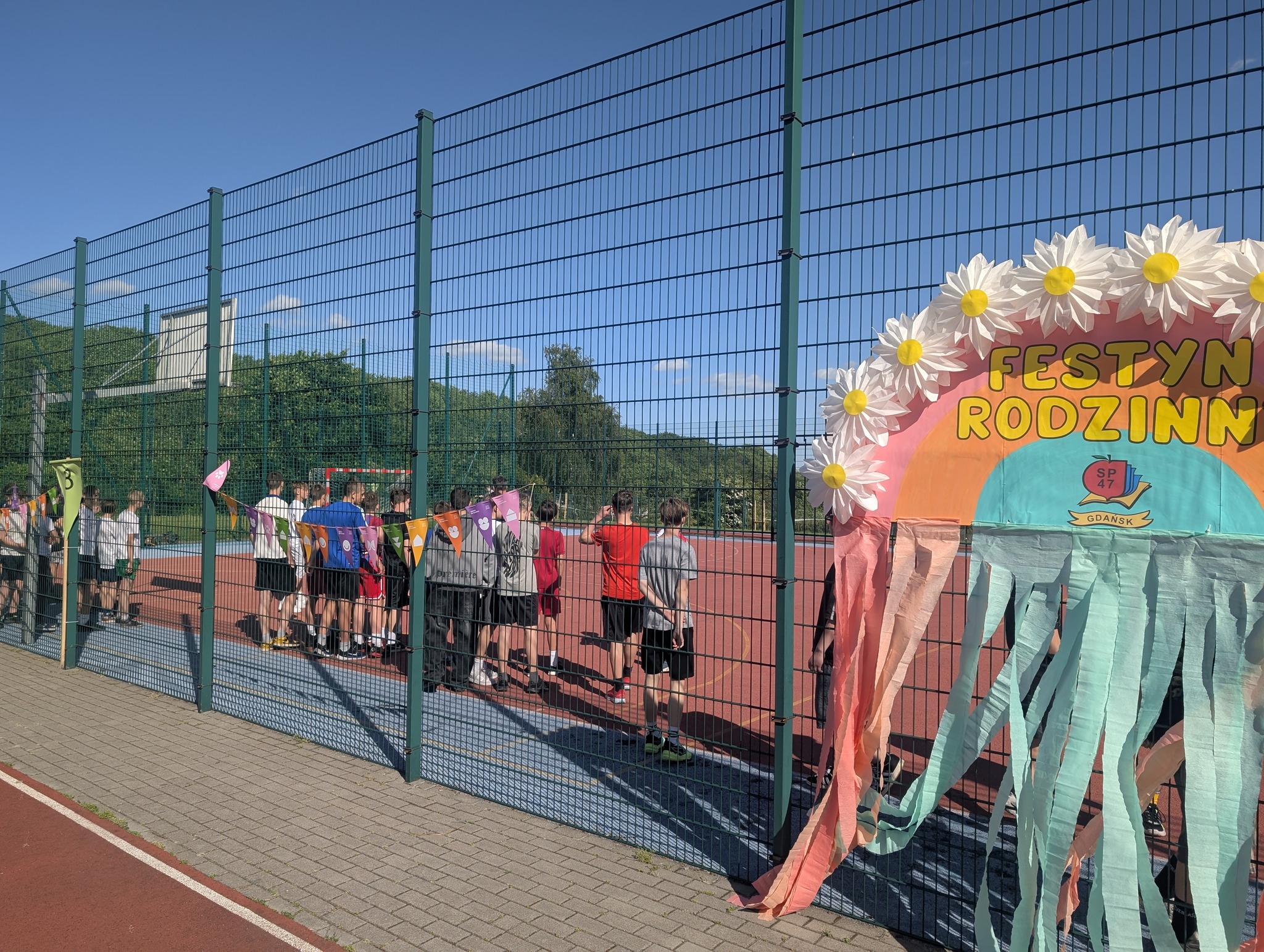 Na pierwszym planie kolorowy baner z podpisem Festyn Rodzinny i logo szkoły. W tle ogrodzone boisko szkolne na którym stoją uczniowie.