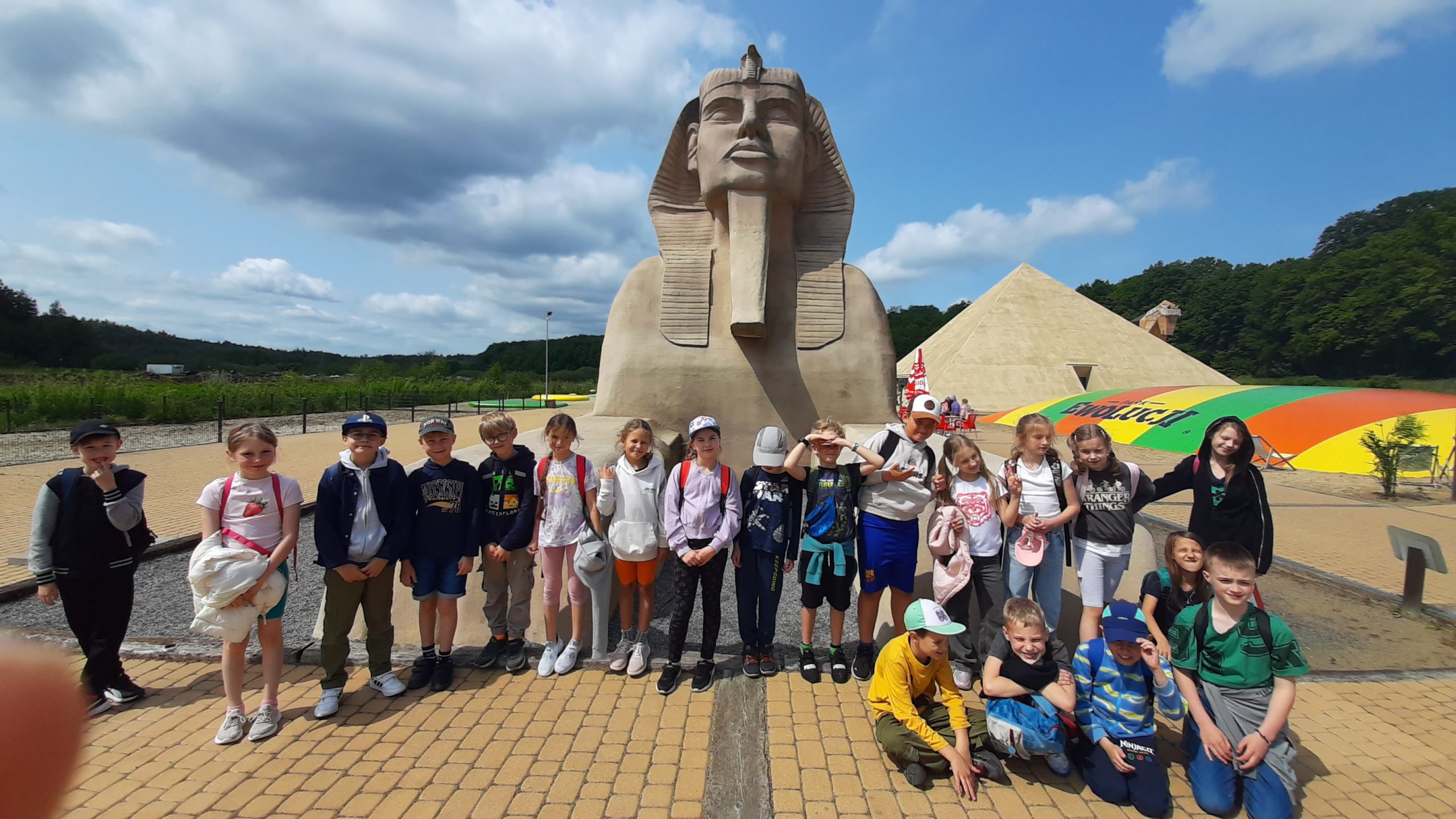 Zdjęcie w plenerze. Grupa dzieci przed makietą Sfinxa stoi w szeregu. Przed nimi siedzi czworo chłopców. W tle po prawej piramida i dmuchaniec. W dali pas drzew.