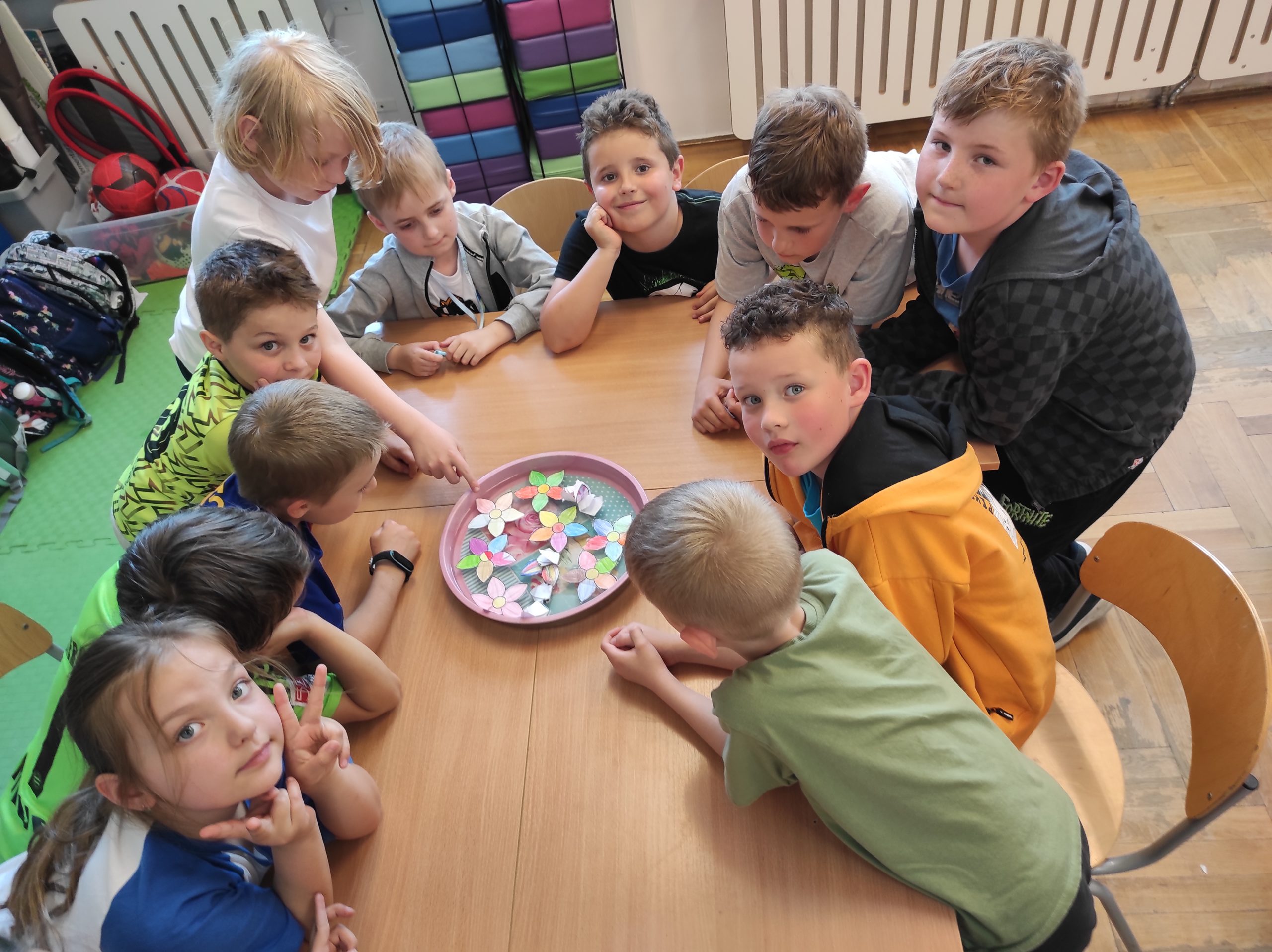 11 dzieci siedzi wokoło złączonych stołów. Na środku leży taca, a w niej widoczne są kwiaty z papieru. Kilka z nich jest częściowo zwinięta, reszta rozwinięta. W tle elementy sali.