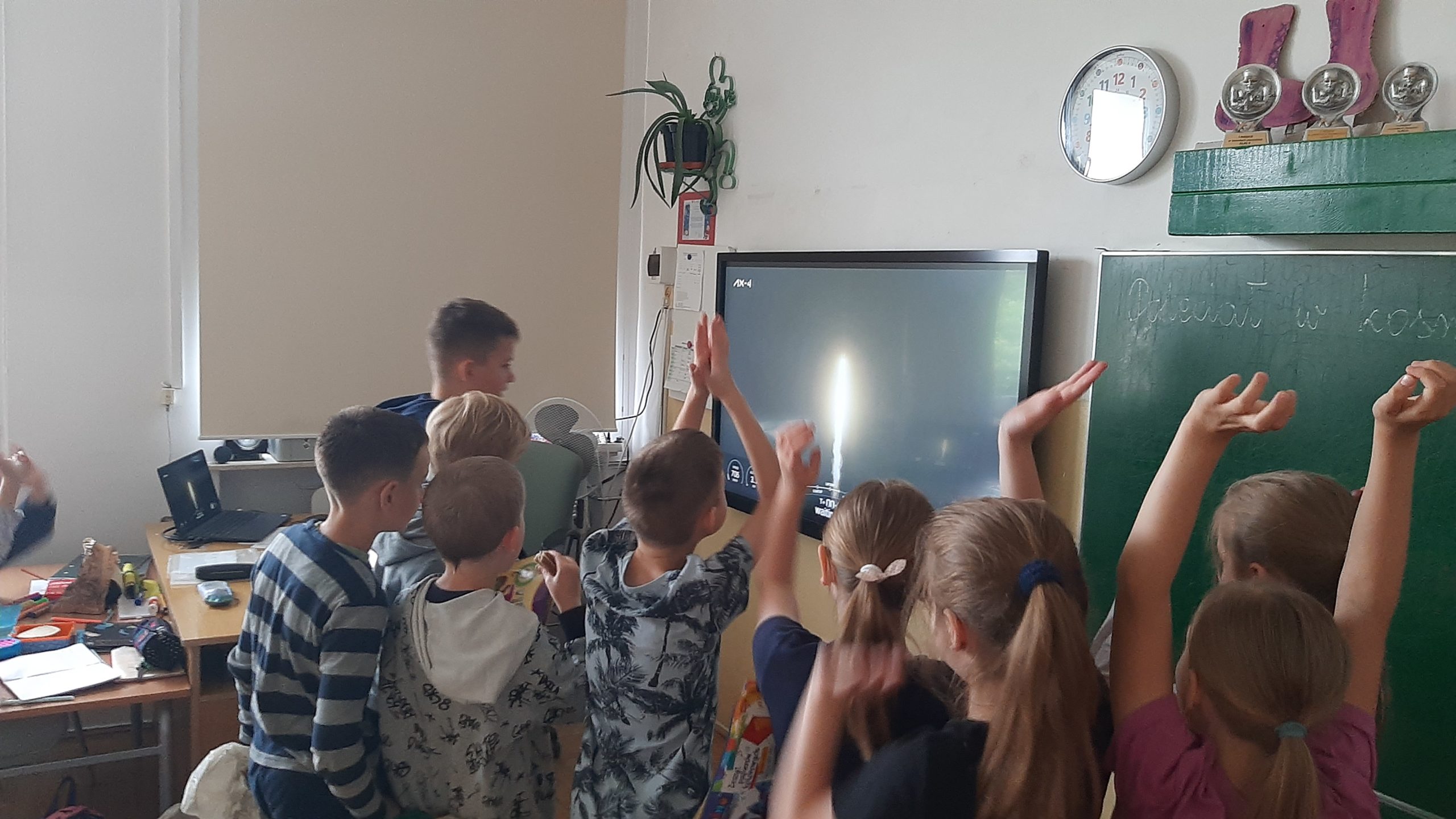 Grupa dzieci stoi przed tablicą interaktywną i patrzy w ekran, na którym widać lecącą w górę rakietę. Niektóre dzieci w geście radości unosi ręce do góry. Na prawo od ekranu tablica z niepełnym napisem: „Poleciał w kosmos.”. Powyżej kinkiet z kwiatem, zegar i półka ze statuetkami. Po lewej zasłonięte roletą okno.