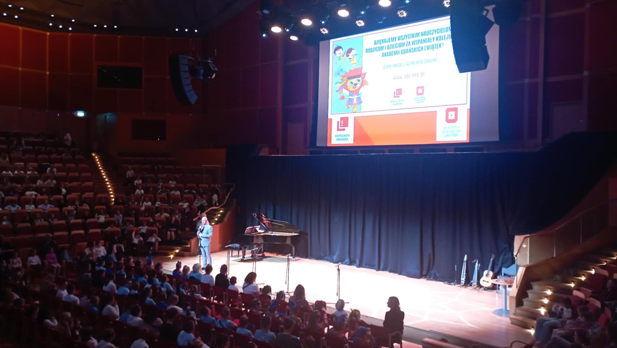 Na scenie Filharmonii Bałtyckiej przemawia Pomorski Kurator Oświaty. Nad sceną na telebimie wyświetlają się napisy dotyczące Akademii Gdańskich Lwiątek. Na krzesłach w wielu sektorach zasiadają widzowie.