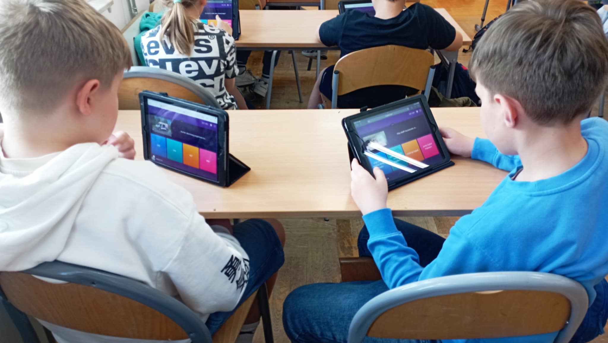 W szkolnej ławce na krzesłach dwaj chłopcy obróceni plecami. Na ławkach IPady wyświetlające quiz na Quizizz. W tle inni uczniowie w ławce.