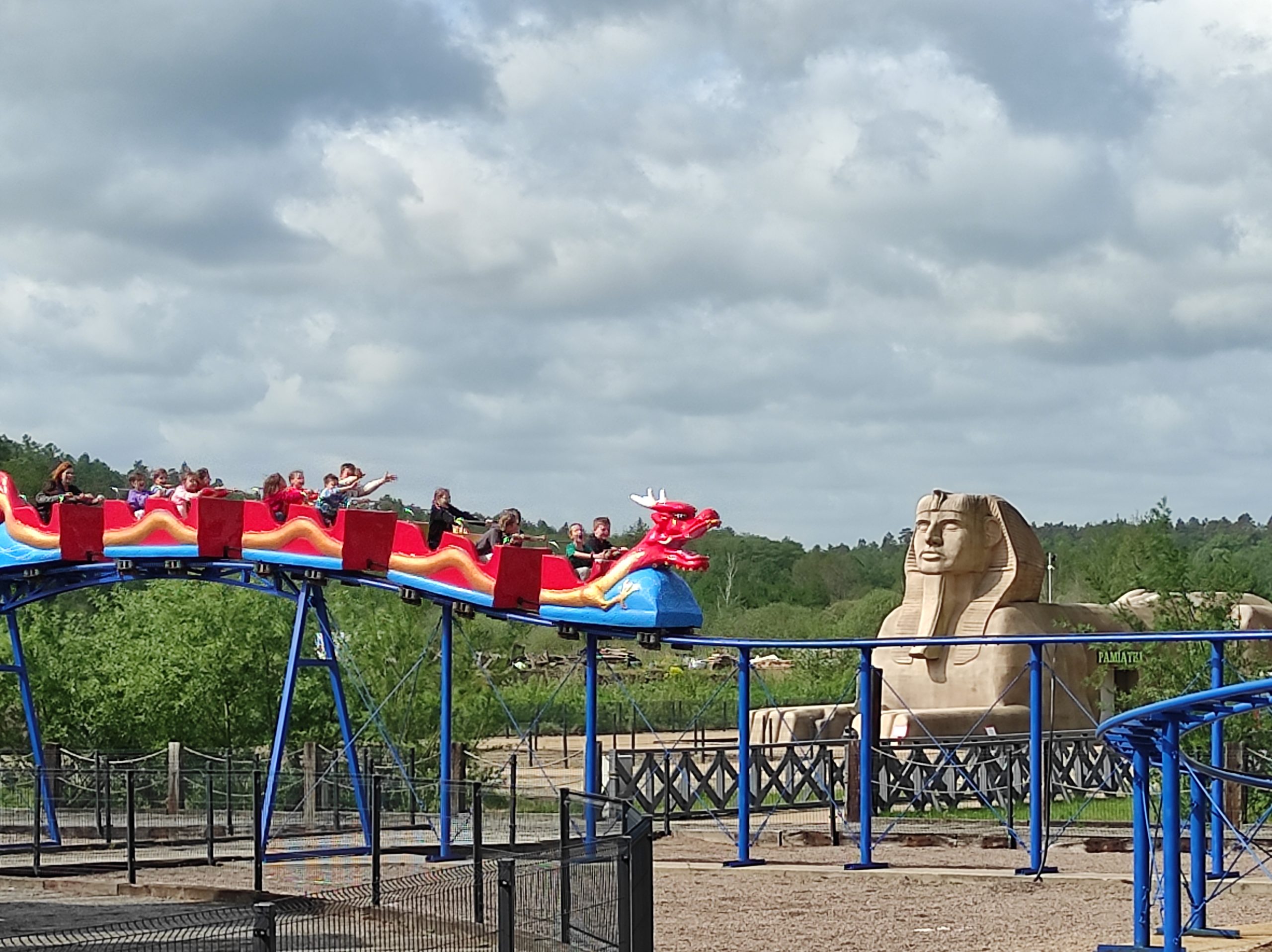 Zdjęcie w plenerze. Tor saneczkowy, a na nim wagoniki, pierwszy z nich z motywem smoka. W wagonikach siedzą siedzi. W tle duży  Sfinx.