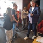 Uczniowie z klasy 8A stoją w kolejce po odbiór medali i trofeów. Dorosły mężczyzna w okula-rach i granatowej marynarce rozdaje medale, trzymając je w ręku. Jeden z chłopców trzyma złoty puchar, a pozostali uczestnicy mają zawieszone medale na szyjach.