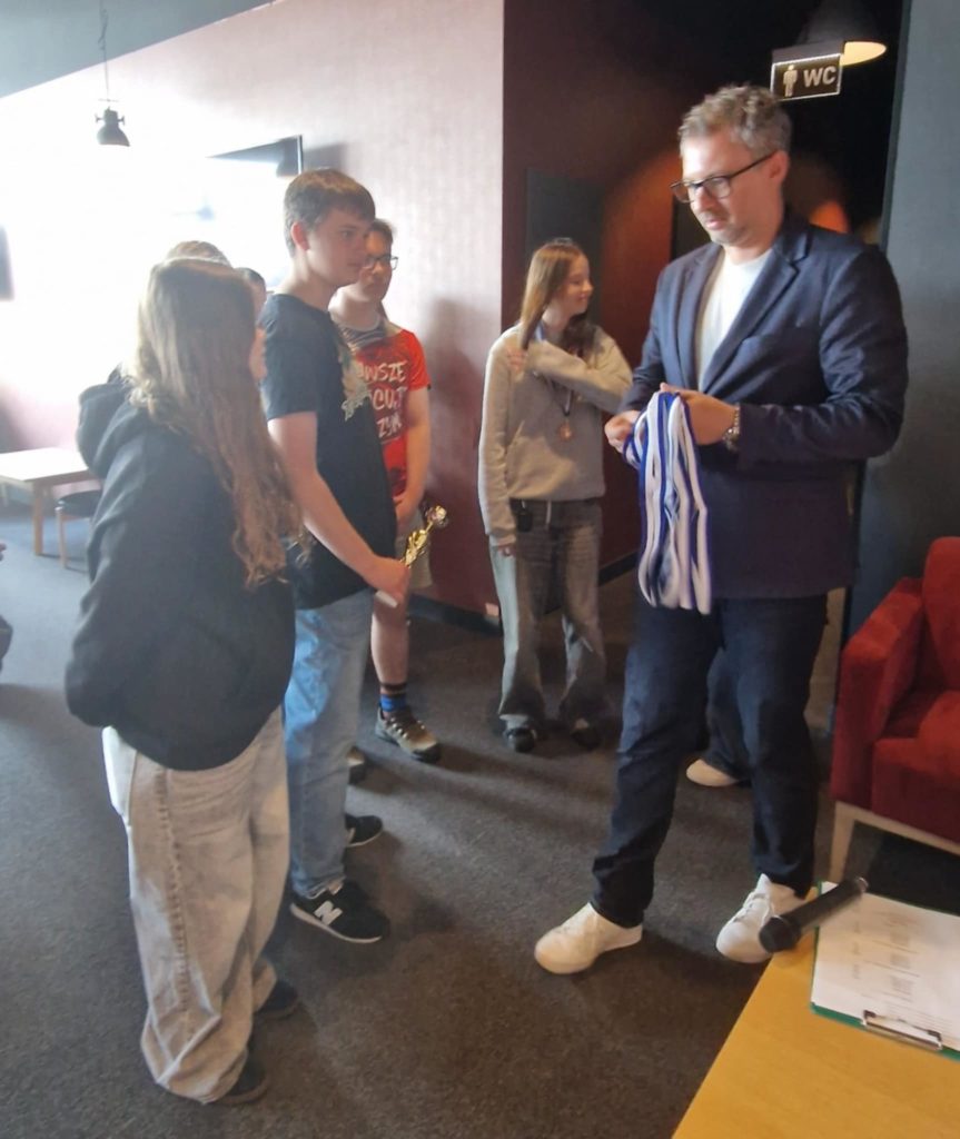 Uczniowie z klasy 8A stoją w kolejce po odbiór medali i trofeów. Dorosły mężczyzna w okula-rach i granatowej marynarce rozdaje medale, trzymając je w ręku. Jeden z chłopców trzyma złoty puchar, a pozostali uczestnicy mają zawieszone medale na szyjach.