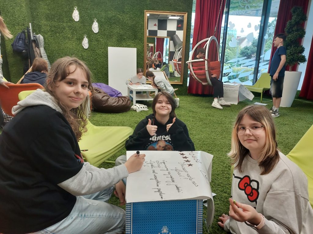 Trzy dziewczyny pracują wspólnie przy niskim stoliku, na którym leży duży arkusz papieru z zapisanymi pomysłami i rysunkami. Jedna z dziewczyn pokazuje uniesione kciuki w geście aprobaty, pozostałe uśmiechają się do aparatu. Wnętrze jest pokryte sztuczną trawą, z kolorowymi fotelami, pufami i charakterystycznymi huśtawkami w tle. Za oknem widoczne są na-klejki w kształcie serc oraz zieleń.