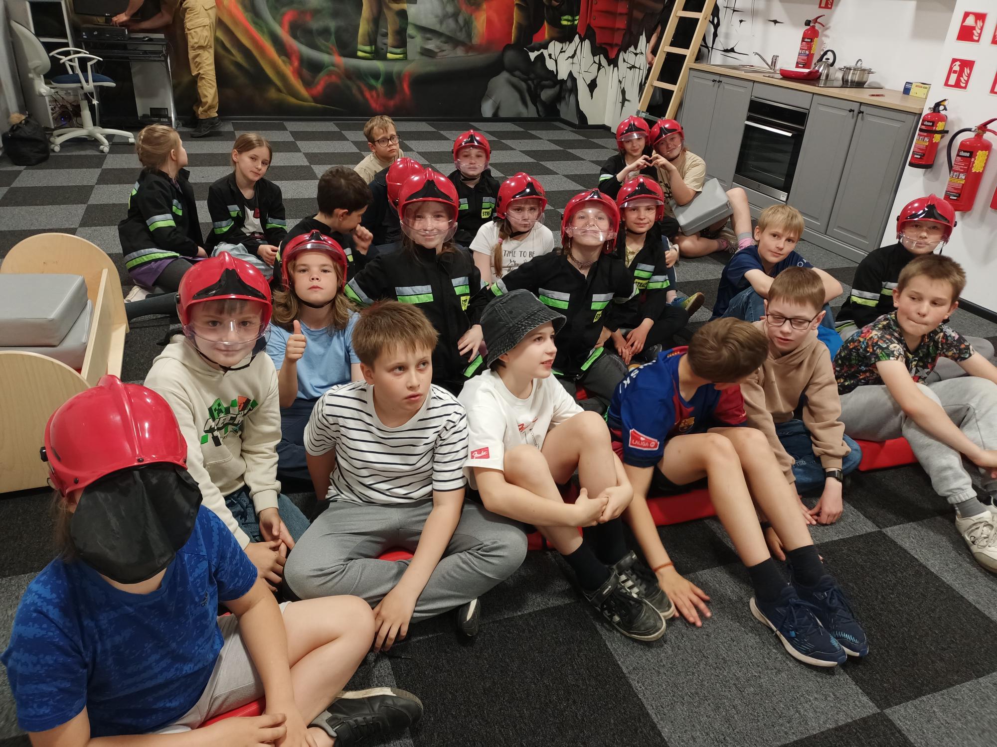 Grupa dwudziestu pięciu uczniów siedzi na dywanie w pomieszczeniu szkoleniowym w Straży Pożarnej. Połowa dzieci jest ubrana w strój strażaka, a na głowach mają czerwone kaski. W tle jest ściana, na której namalowano strażaka gaszącego pożar.