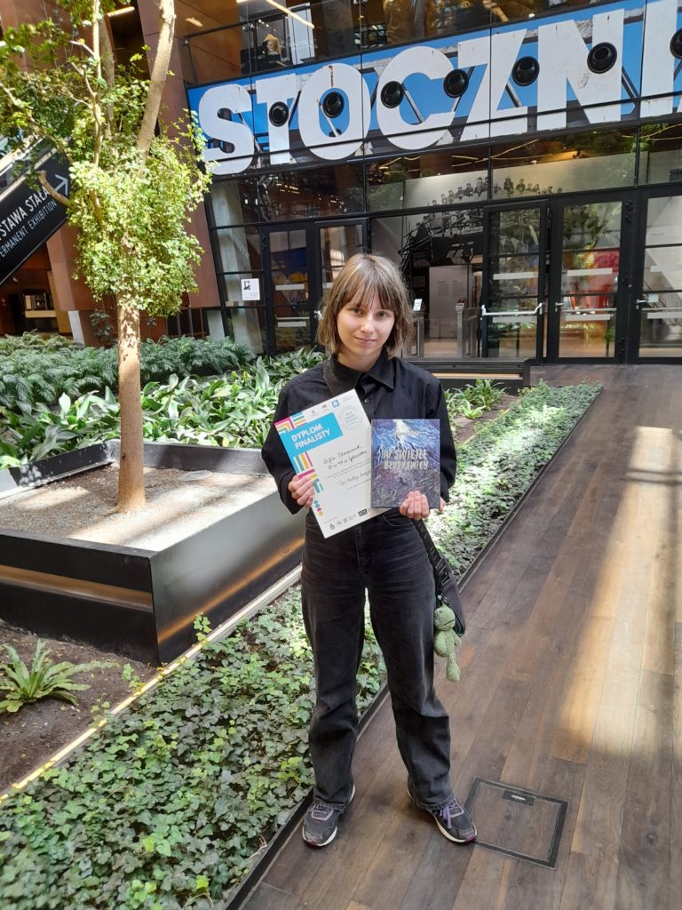 Dziewczynka prezentuje odebrany dyplom na tle napisu „STOCZNIA”