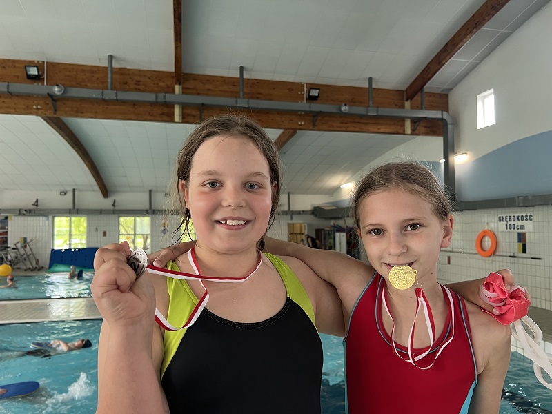 Dwie uczennice z medalami , jedna trzyma go w dłoni a druga zębami, pozuje do zdjęcia