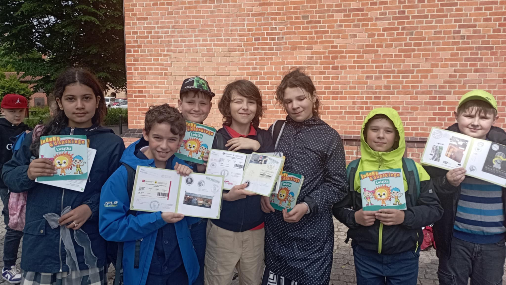 Przed fasadą ceglastego budynku Filharmonii Bałtyckiej stoi ośmioosobowa grupa uczniów klasy 3c z indeksami ALG w dłoniach.