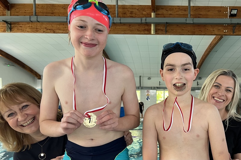 Dwóch uczniów z medalami , jeden trzyma medal dłońmi a drugo zębami , a po bokach twarze dwóch uśmiechniętych pań