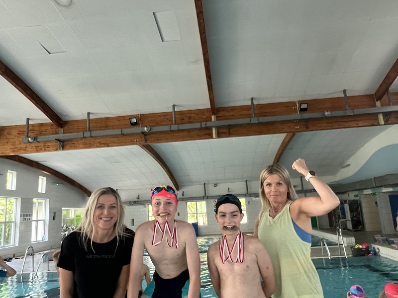 Dwóch uczniów trzyma zębami zdobyte medale a po bokach dwie panie na tle niecek pływackich